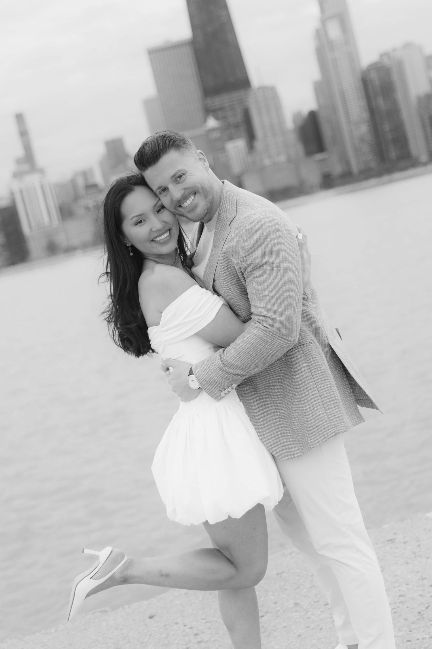 Black and white photo of a couple embracing on a beach with Chicago skyline in the background.