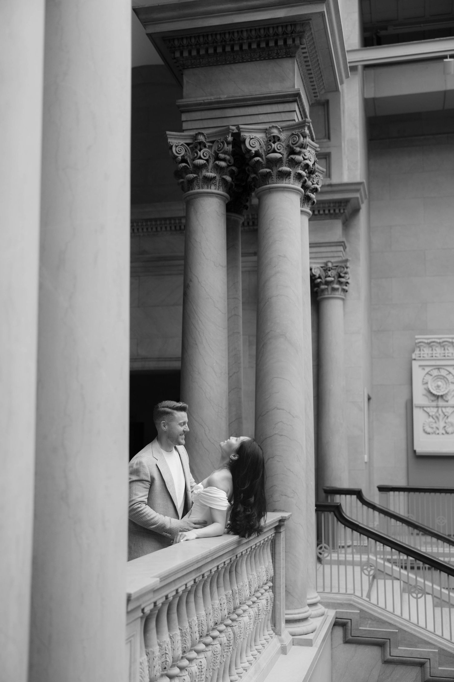 Black and white photo of a couple sharing a romantic moment on a grand balcony with tall classical columns.