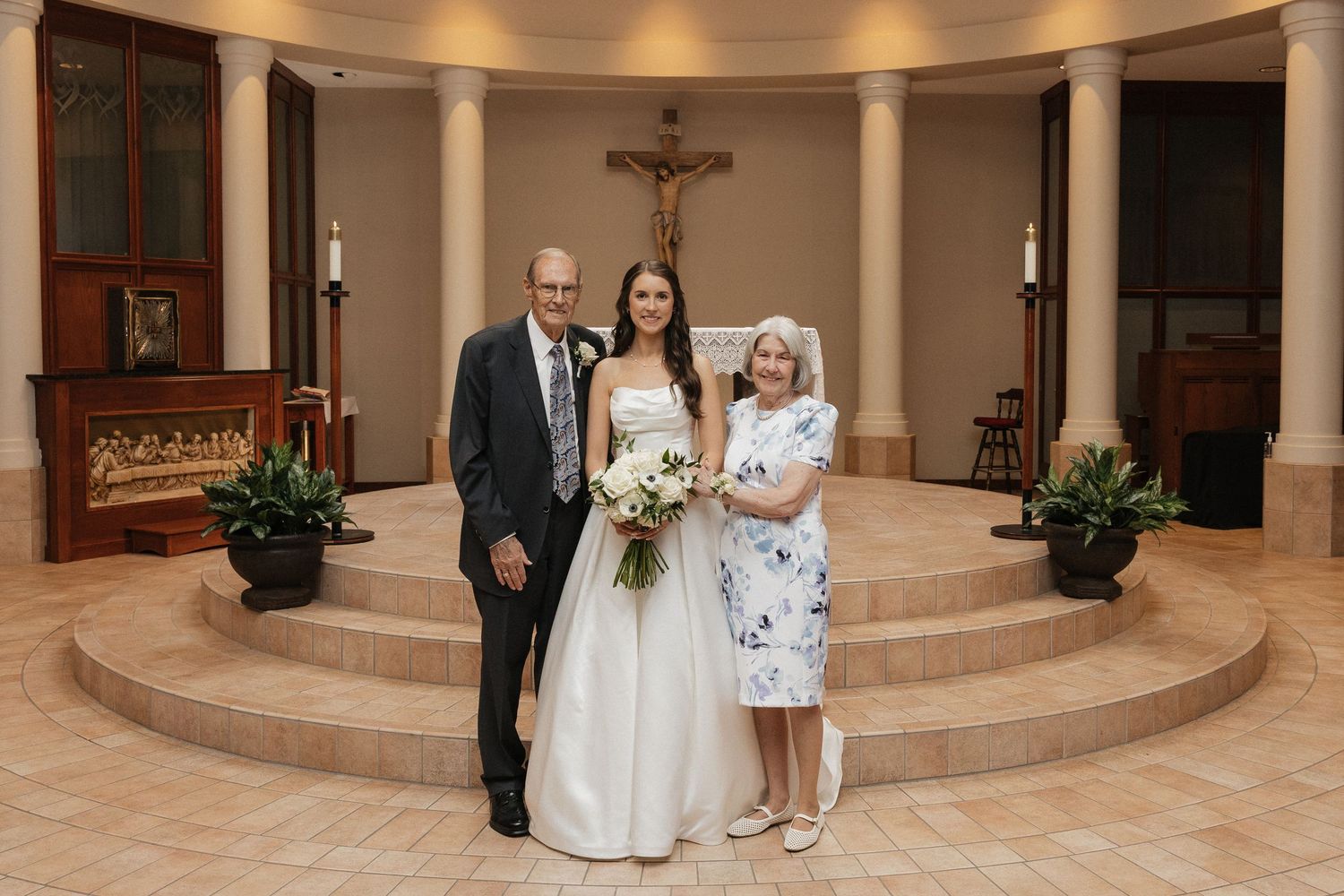 Bride in white gown poses with two guests in a church chapel featuring a crucifix and marble steps.