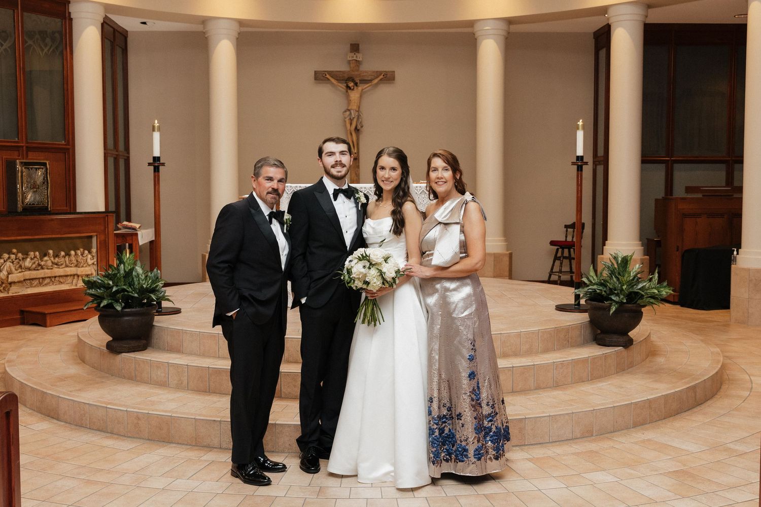 Wedding party poses in front of altar with crucifix, bride in white gown holding bouquet with groom and family.