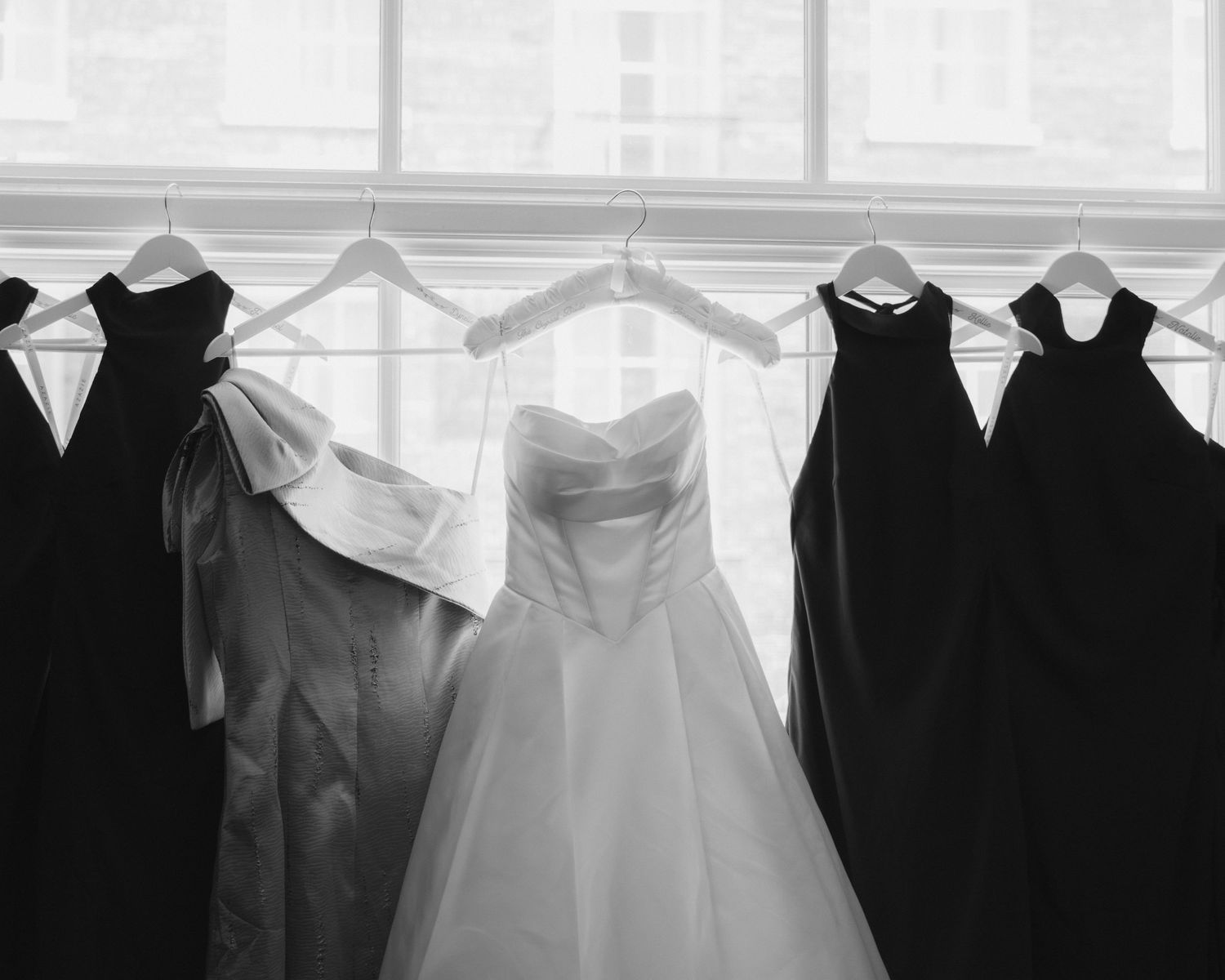 Black and white photo of wedding dress and bridesmaid gowns hanging on window display.