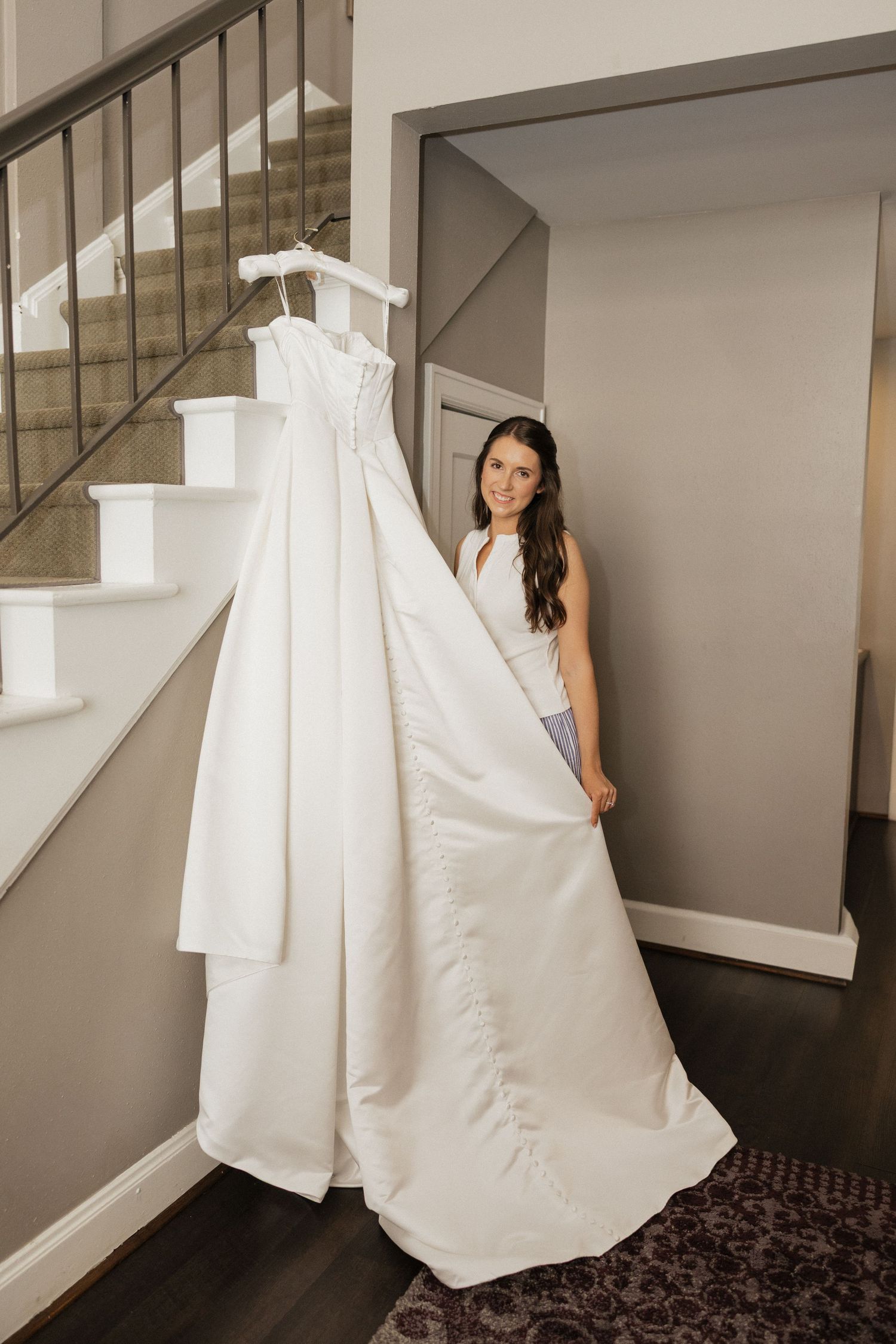 Bride holding up her elegant white wedding gown on a hanger near a staircase inside a home.