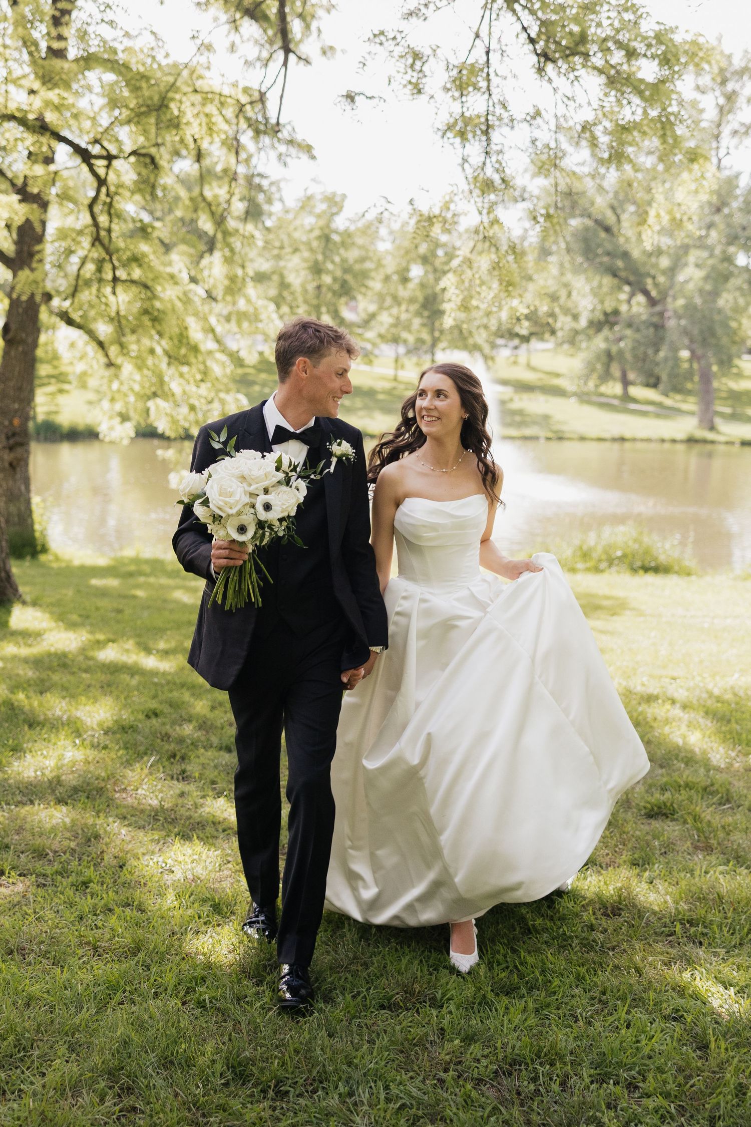 A bride and groom walk hand-in-hand through a lush green park, smiling at each other on their wedding day.