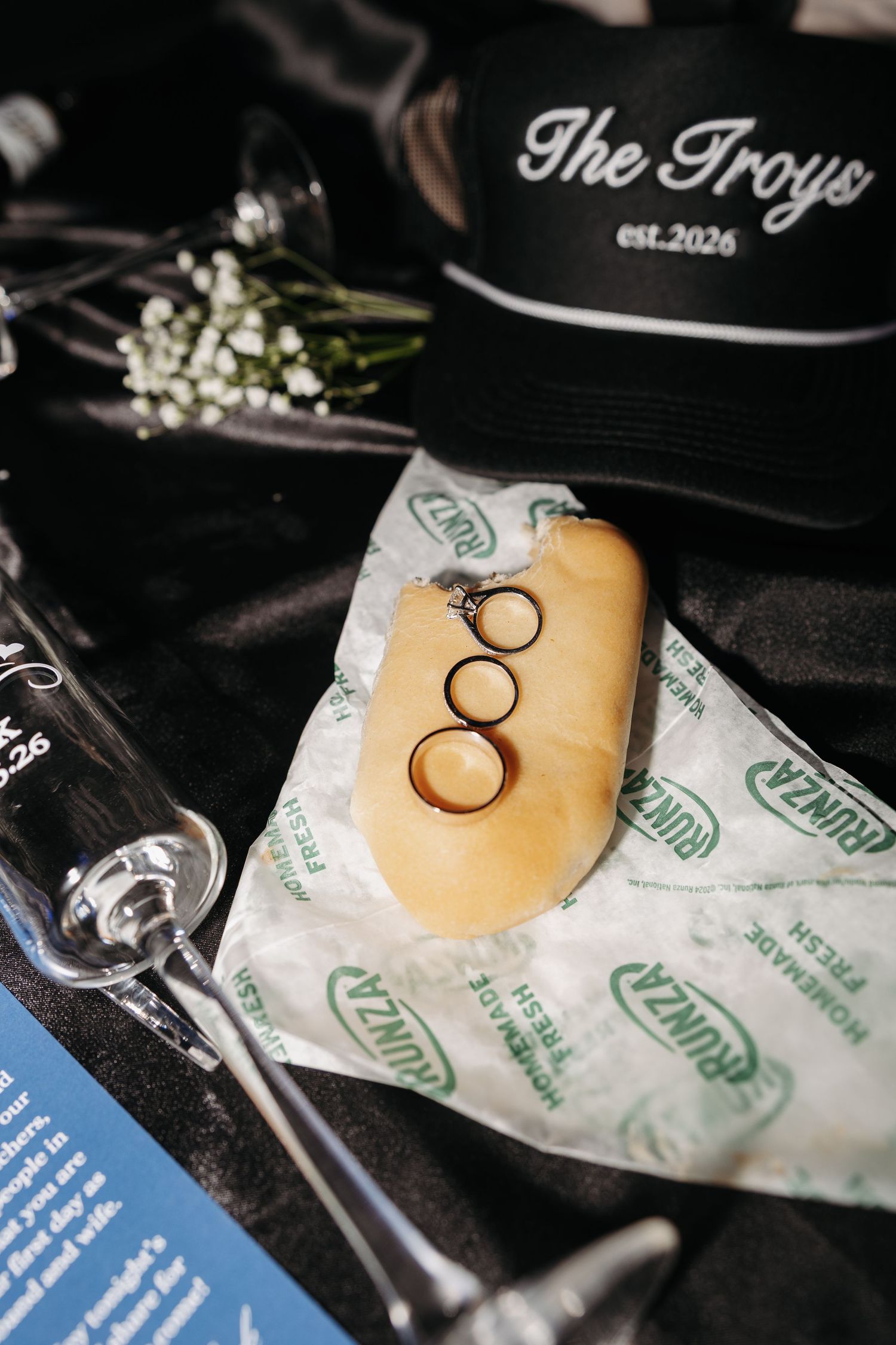 Wedding rings displayed on a Runza sandwich with The Troys est.2026 hat, champagne glass, and flowers on black satin.