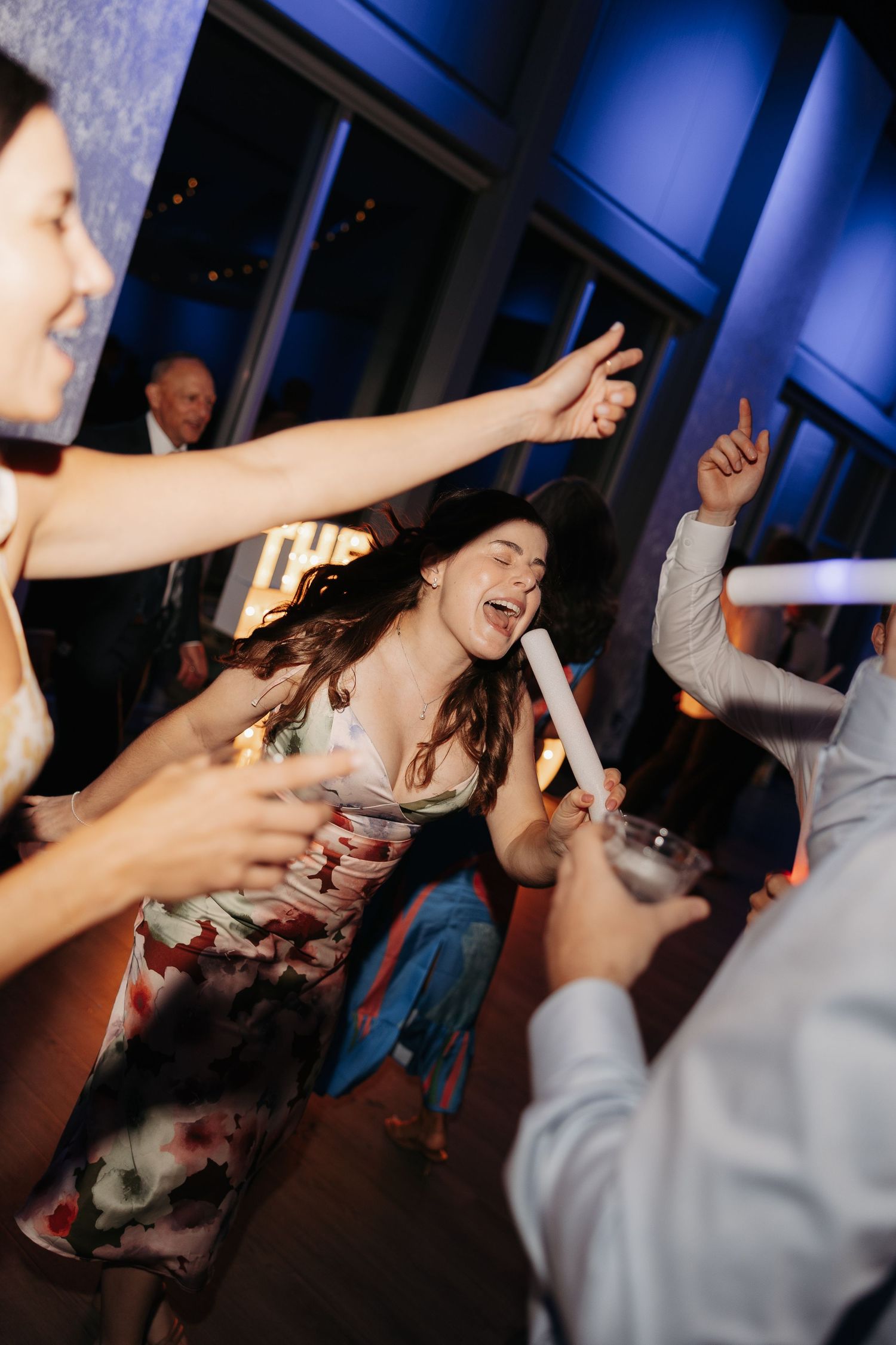 Guests dancing joyfully at a wedding reception, woman in floral dress laughing with glow stick under blue lights.