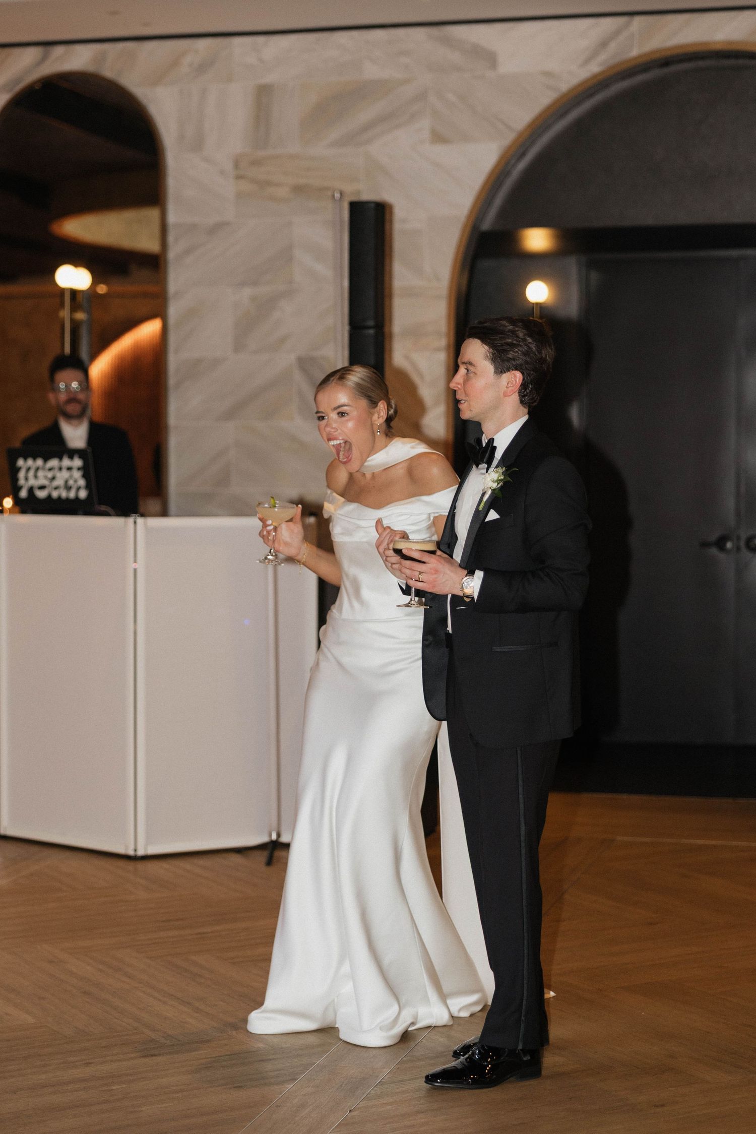 Bride and groom holding champagne glasses and laughing during their wedding reception in an elegant venue.