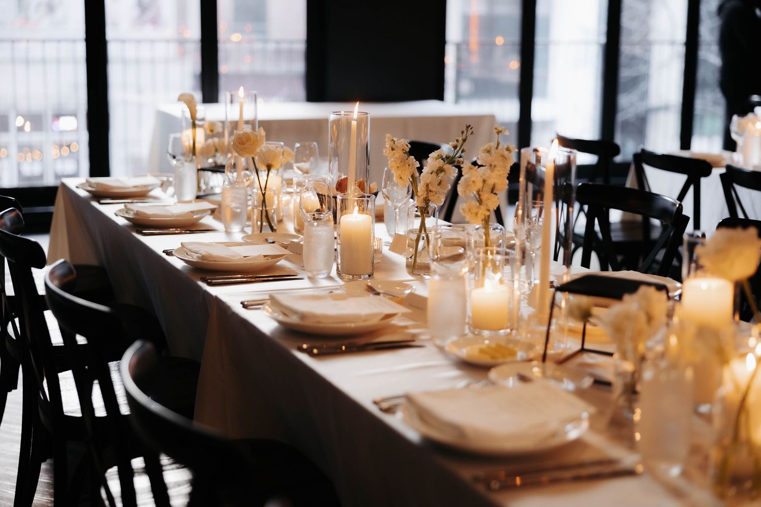 Elegant wedding reception table set with white linens, candles, floral centerpieces, and black chairs in a modern venue.