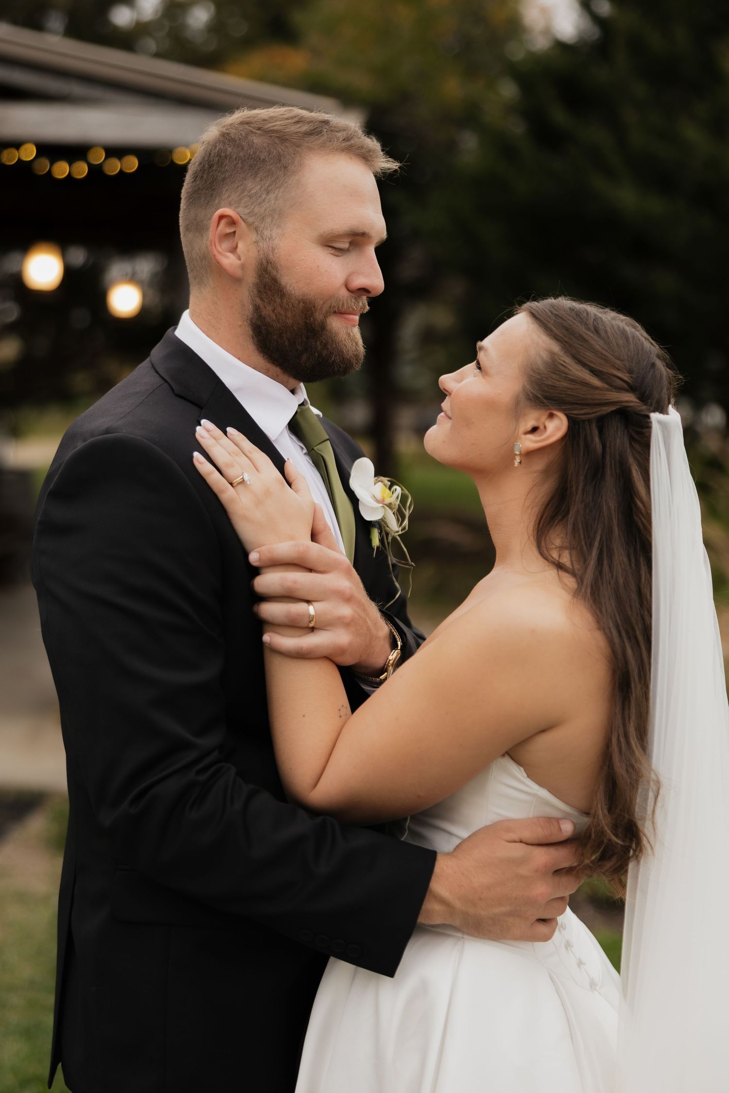 Bride in a white strapless gown and veil embraces a bearded groom in a black suit with a boutonniere during an outdoor wedding.