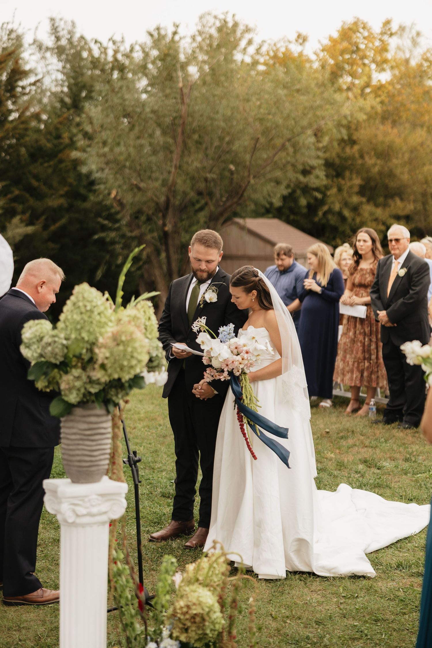 Bride in a white gown and groom in a dark suit exchange vows during an outdoor garden wedding ceremony as guests watch.