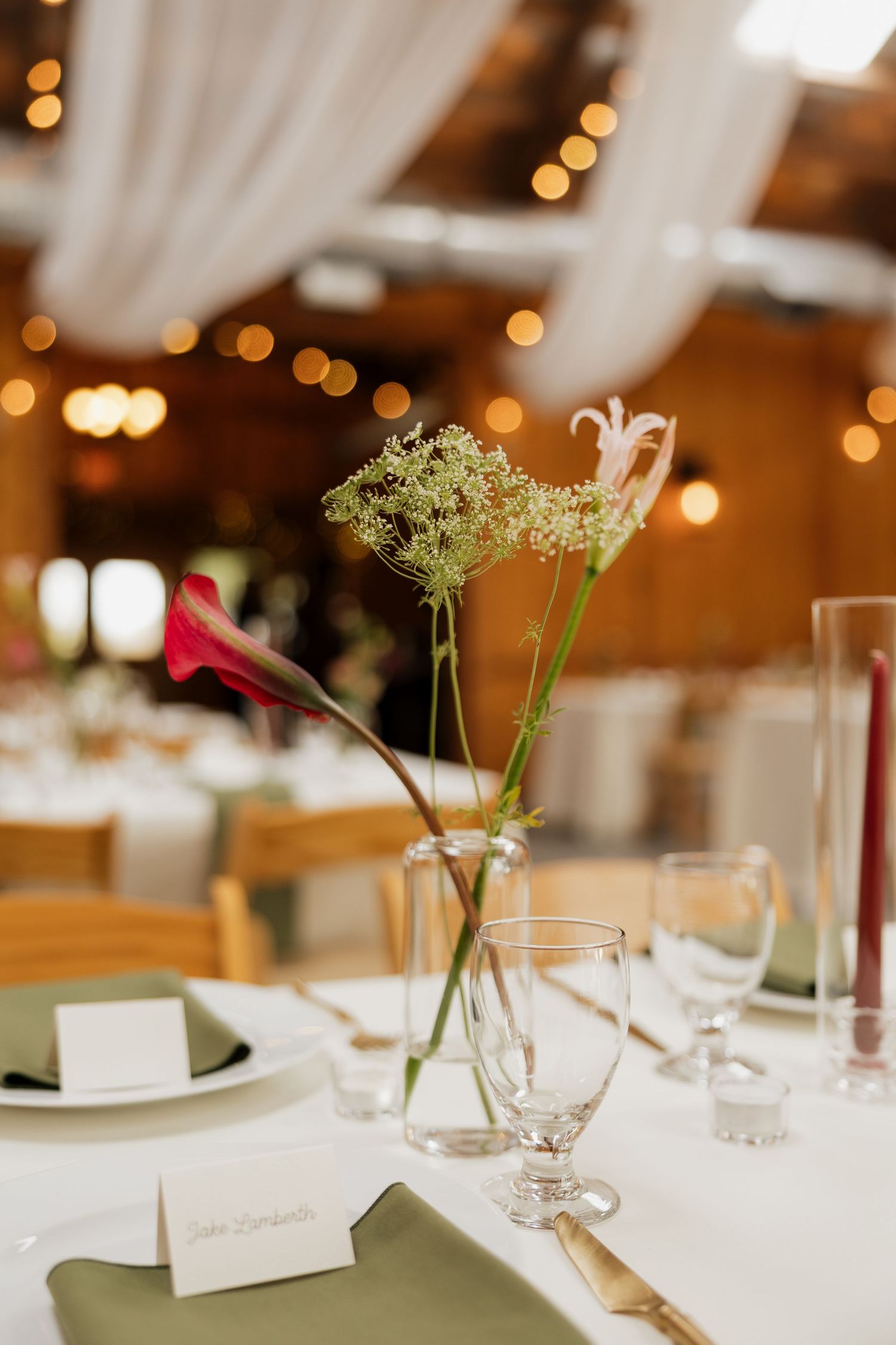 Elegant wedding table setting with olive green napkins, place cards, glassware, and a bud vase of wildflowers under warm string lights.