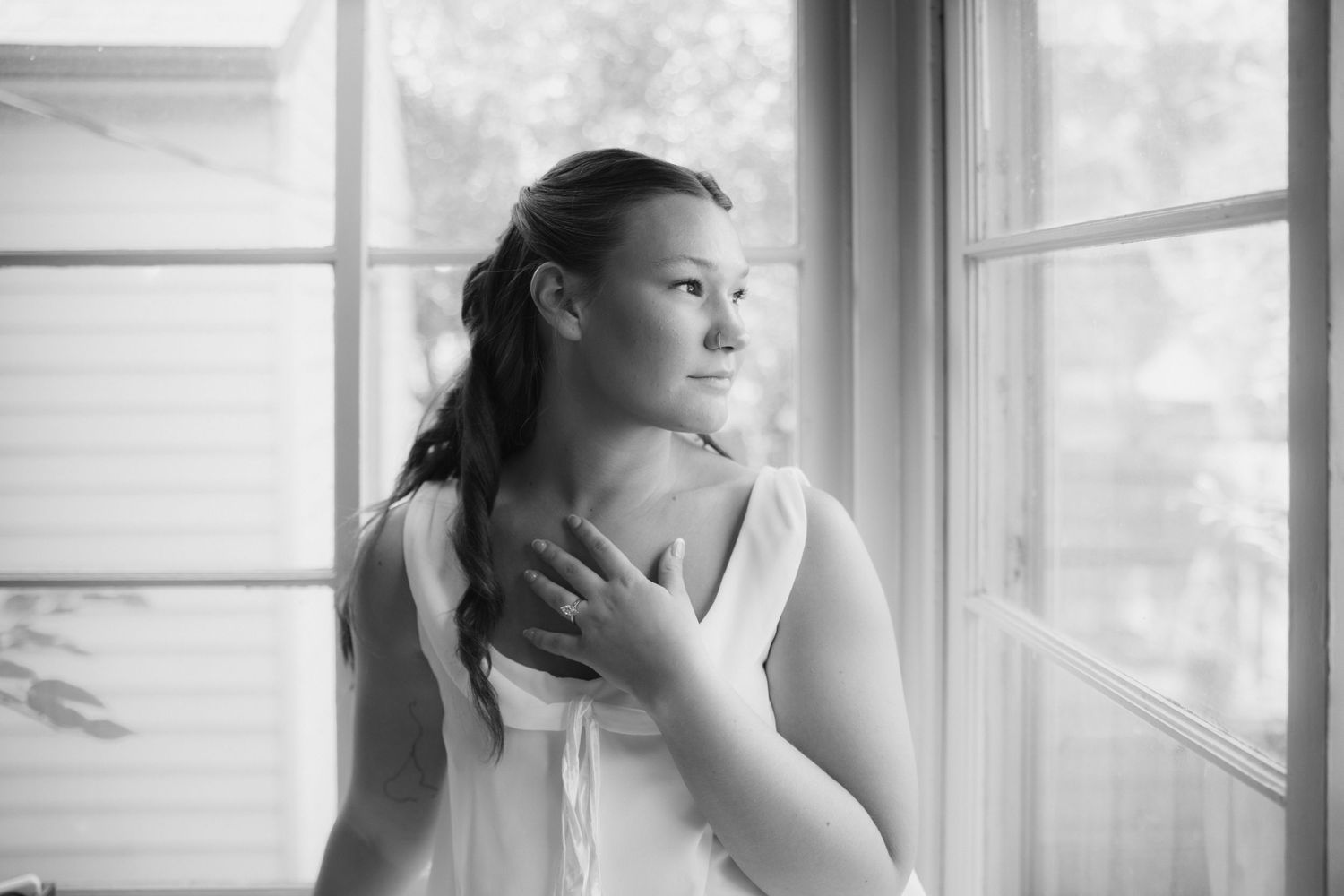 Black and white portrait of a young woman with a ponytail gazing out a window, one hand resting on her chest in soft natural light.