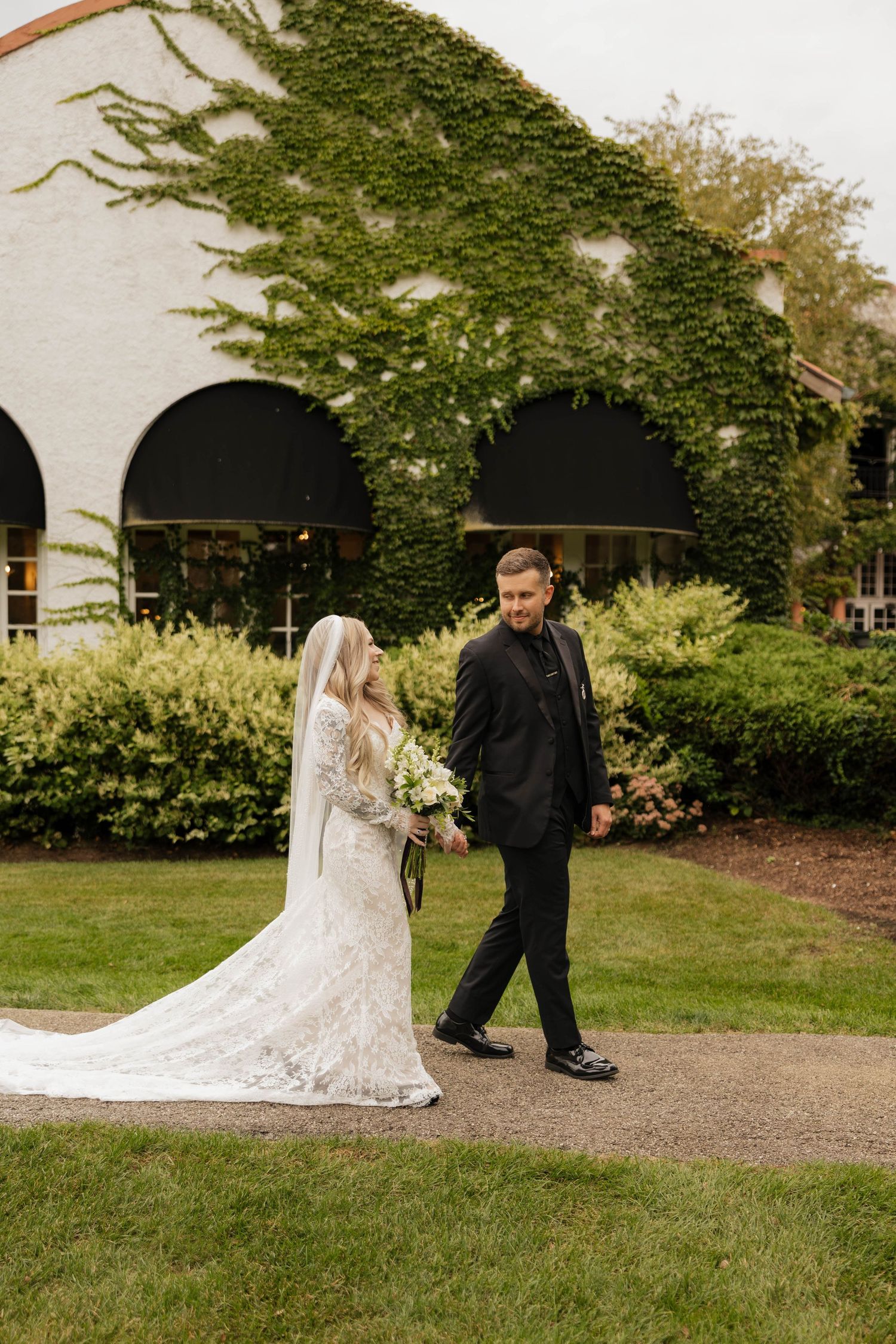 Bride in a lace wedding gown with long veil holds a bouquet and walks hand in hand with a groom in black beside an ivy-covered building.