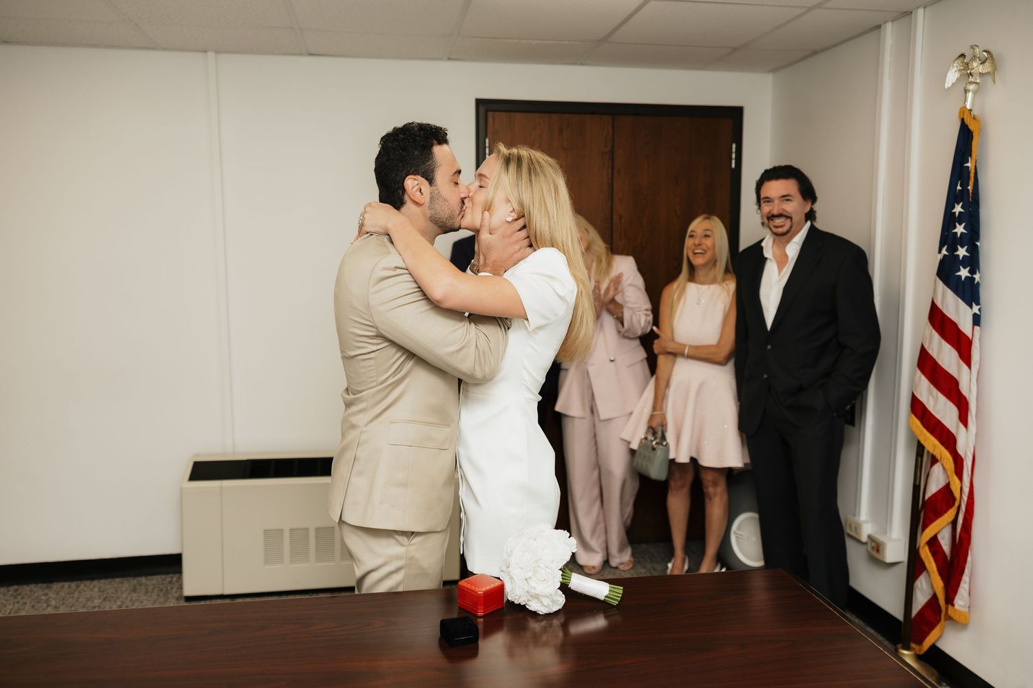 Couple shares a kiss during a civil wedding ceremony in an office with guests and an American flag nearby.