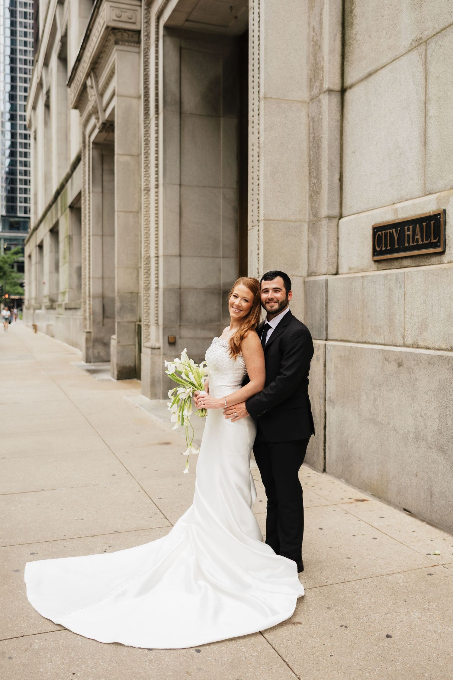 Bride and groom posing outside City Hall, bride in elegant white gown holding bouquet, groom in black tuxedo.