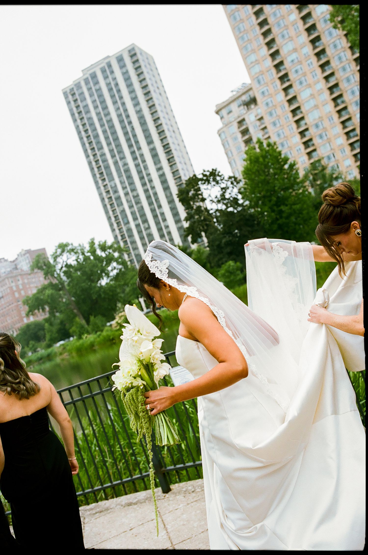 Bride in white gown and veil holds bouquet as bridesmaid adjusts train, city skyline backdrop.
