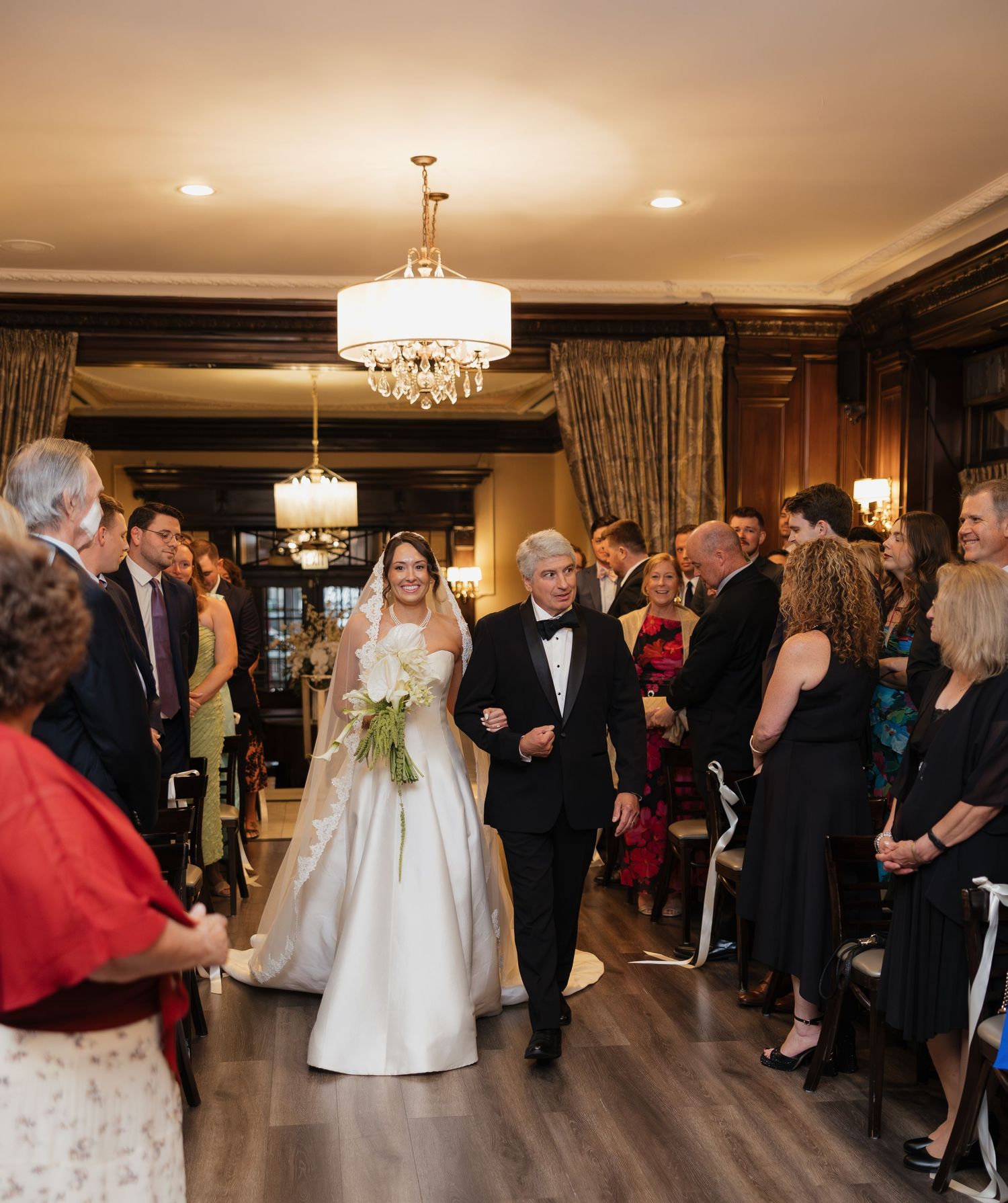 Bride in white gown walks down aisle with escort at elegant indoor wedding ceremony with chandelier lighting.