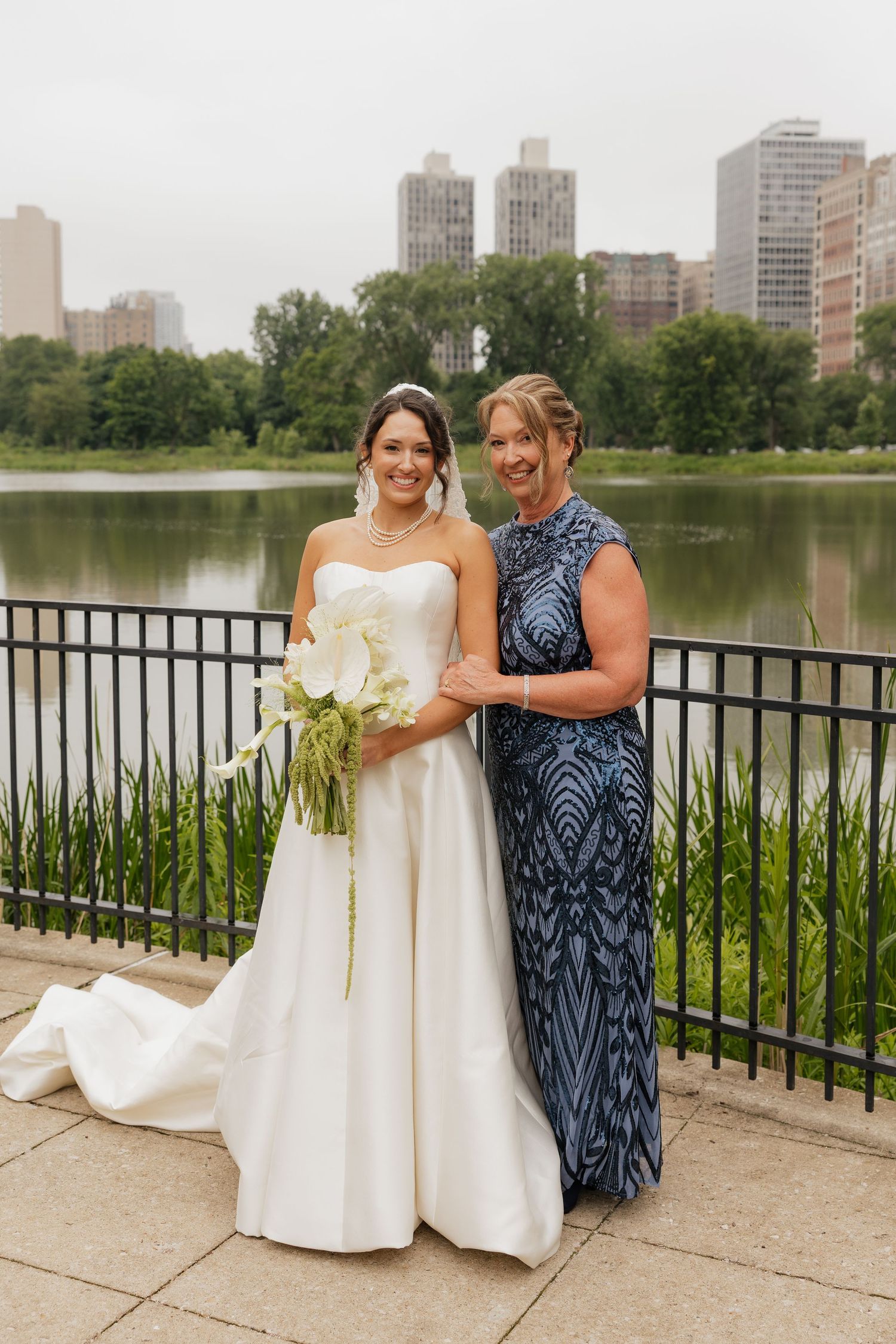Bride in white gown holding bouquet poses with woman in blue sequin dress by lakeside with city skyline backdrop.