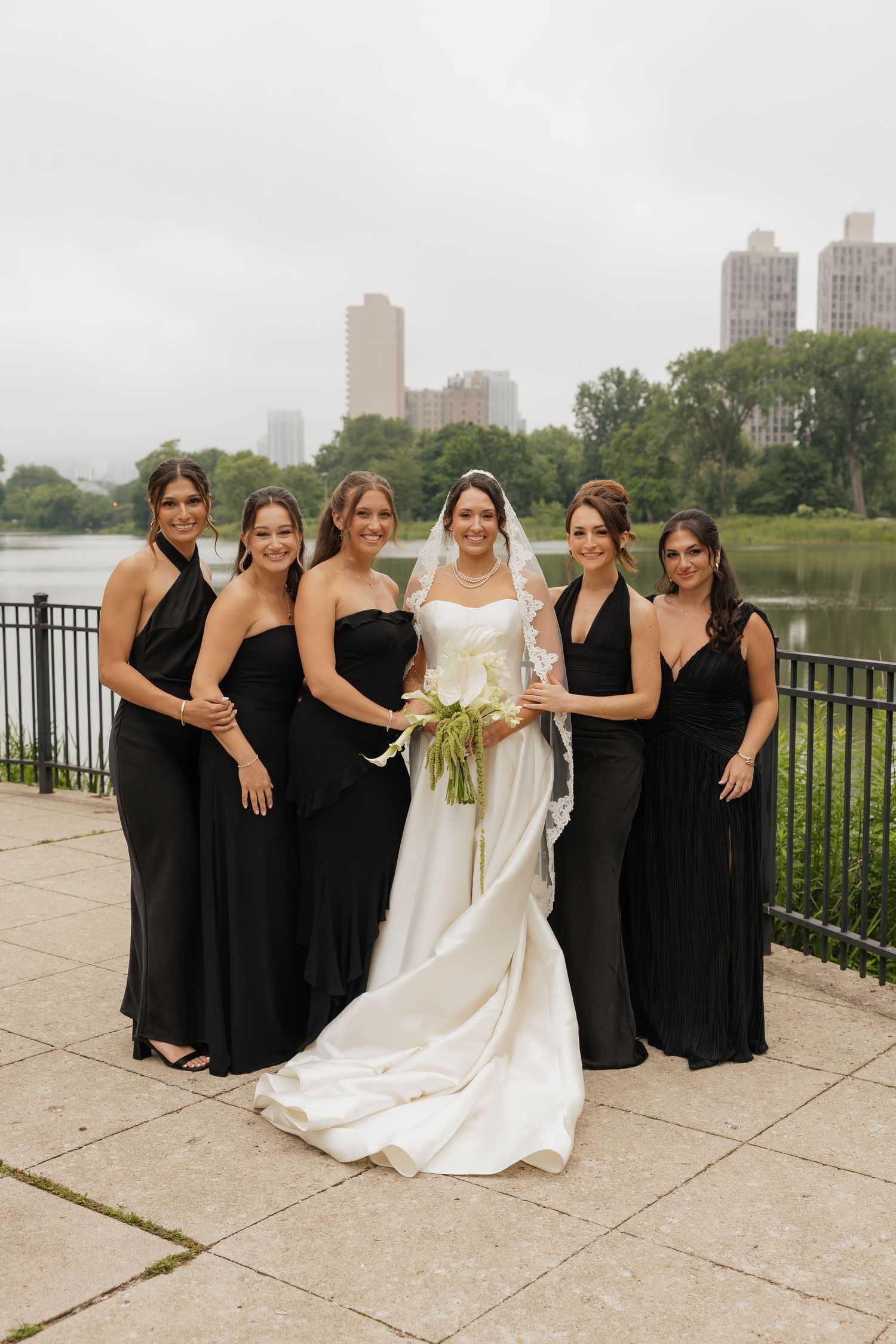 Bride in white gown with bridesmaids in black dresses posing by a lake with city skyline backdrop.