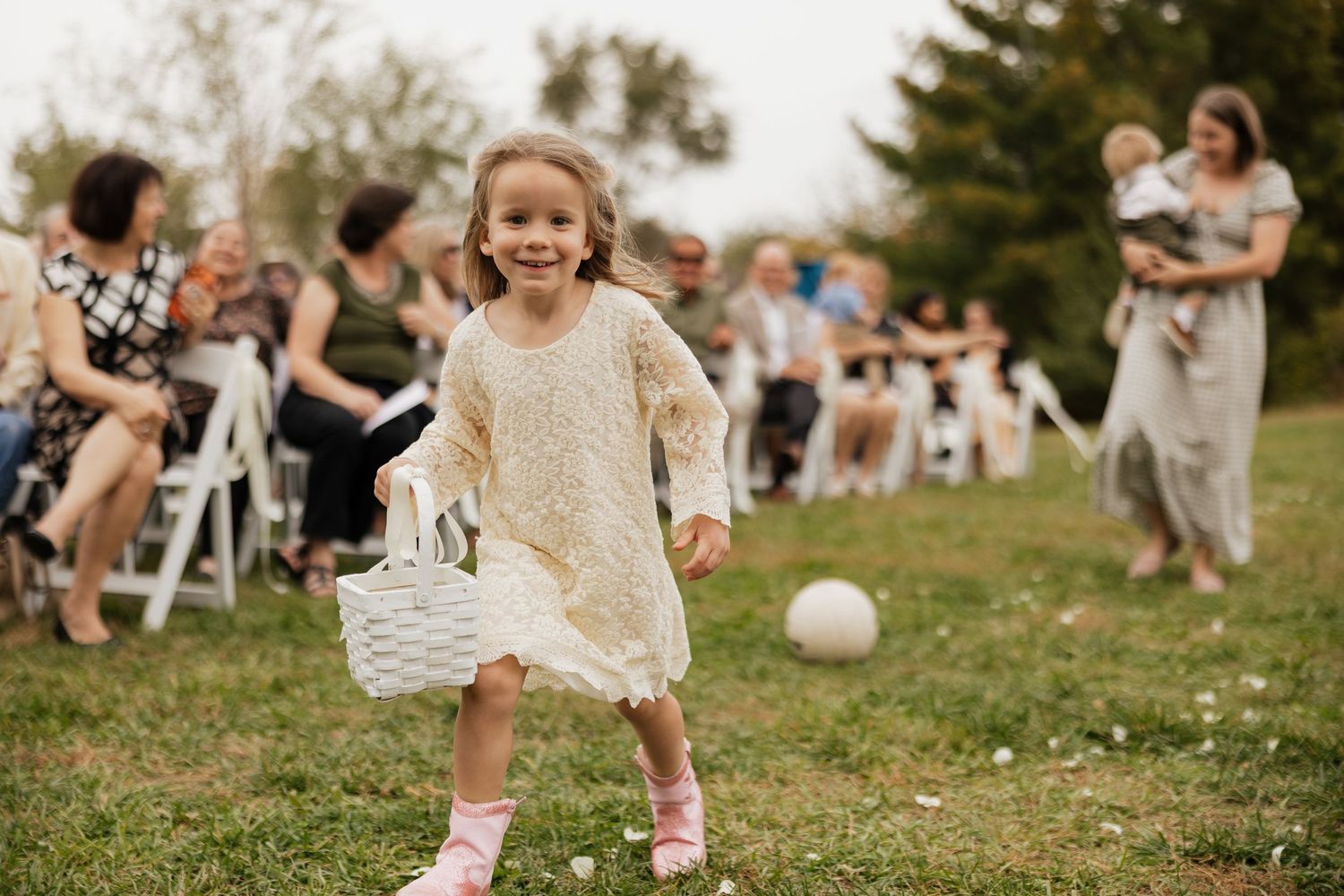 Young flower girl in a cream lace dress and pink boots runs across the grass carrying a white basket at an outdoor wedding ceremony.