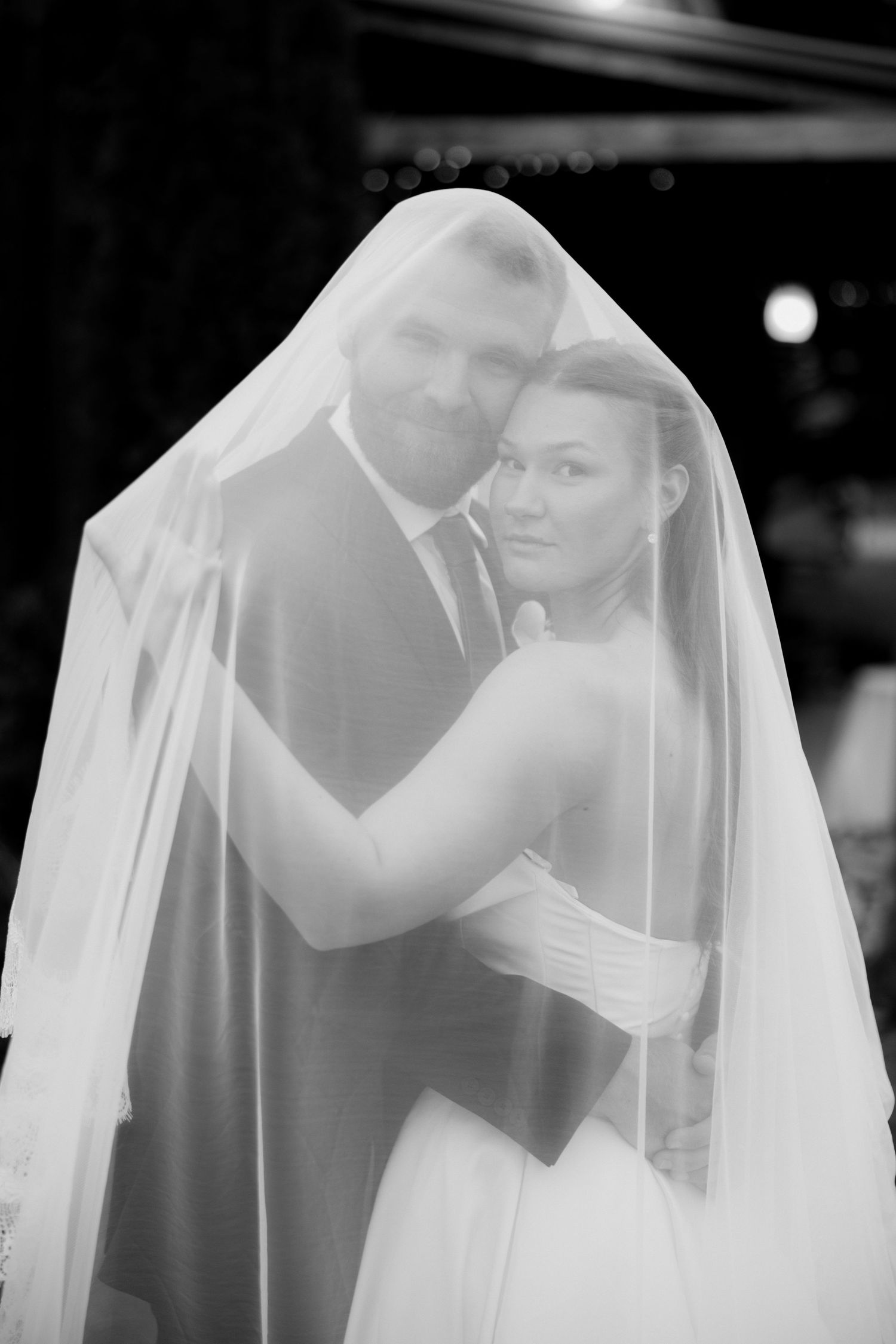 Black and white photo of a bride and groom embracing closely under a sheer wedding veil during their wedding day.