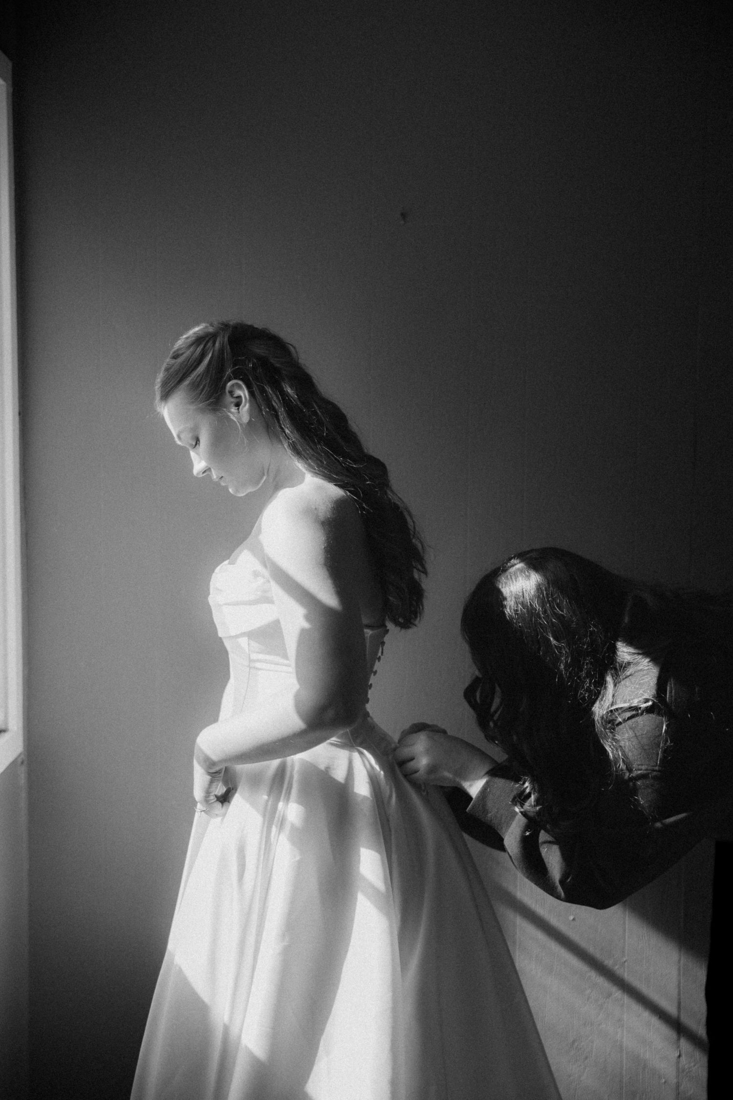 Black and white photo of a bride in a strapless gown standing by a window while a woman fastens the back of her dress.