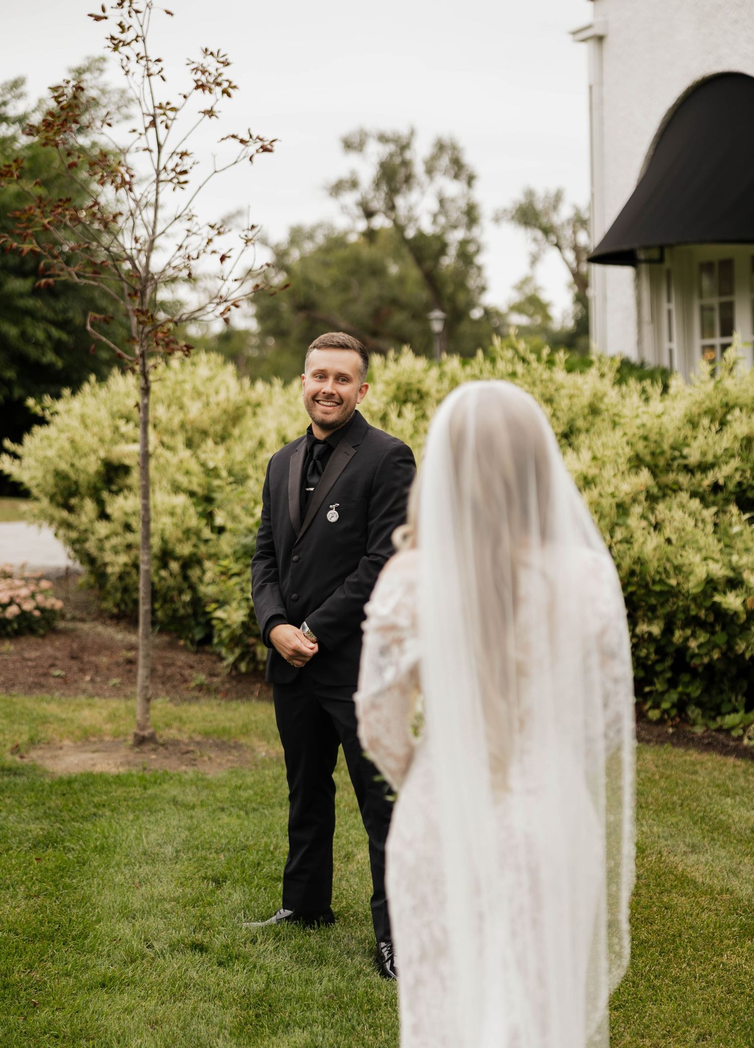 Smiling groom in a black suit watches his bride in a lace gown and long veil during a first look on a sunny garden lawn.