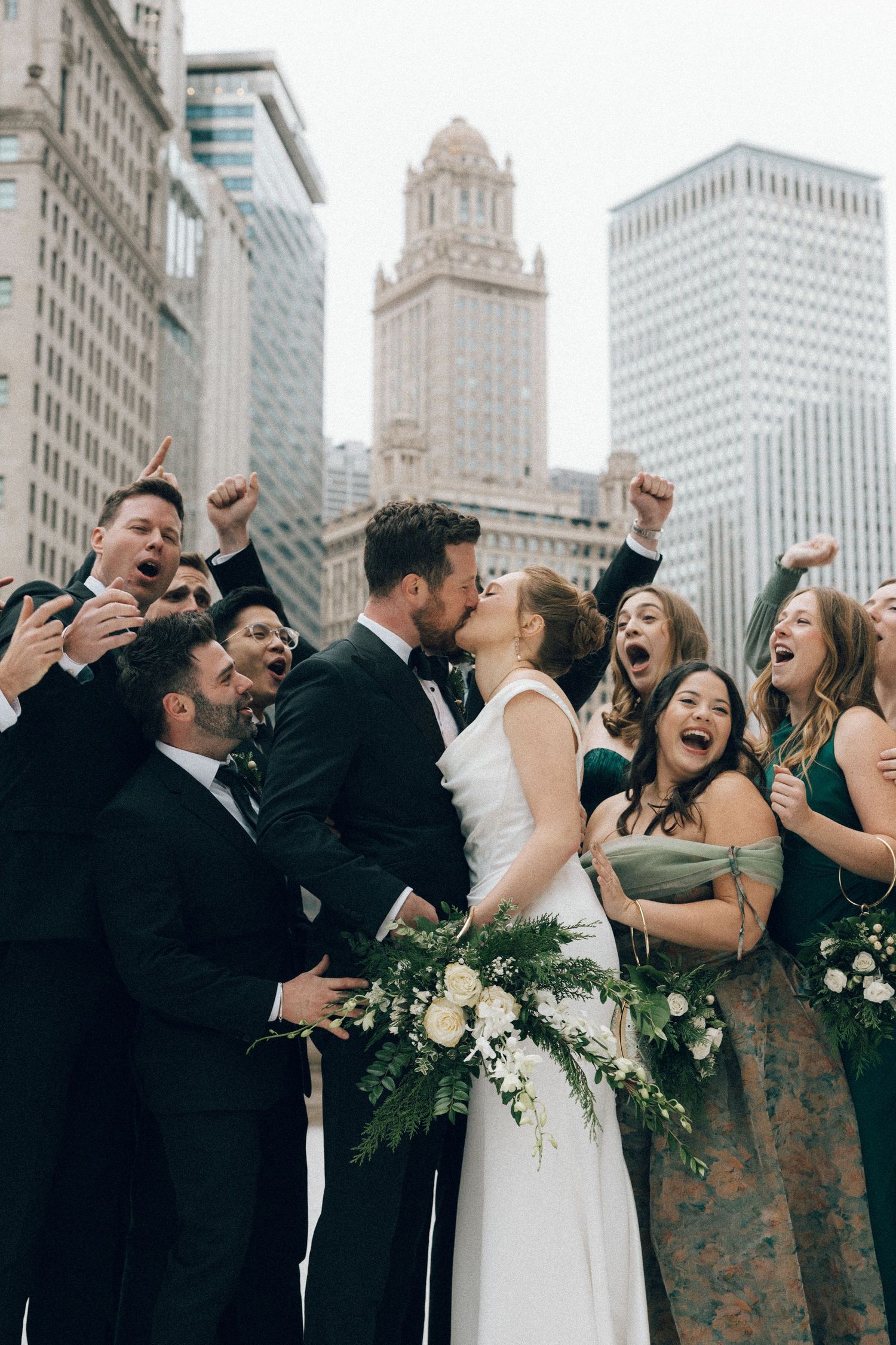 Wedding party laughs joyfully together on rooftop with city skyline in background.