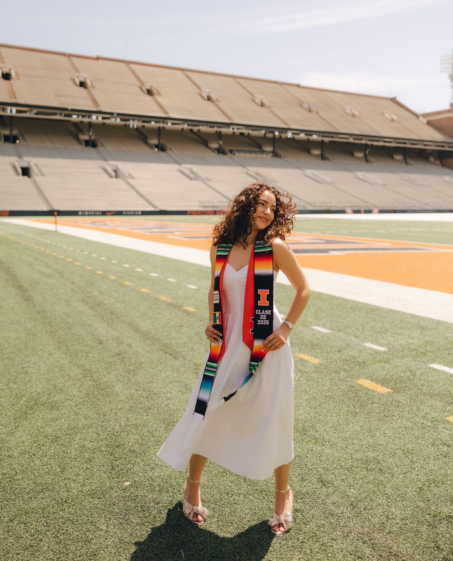 Person wearing a white dress and boots stands on a football field holding a camera in a creative pose.