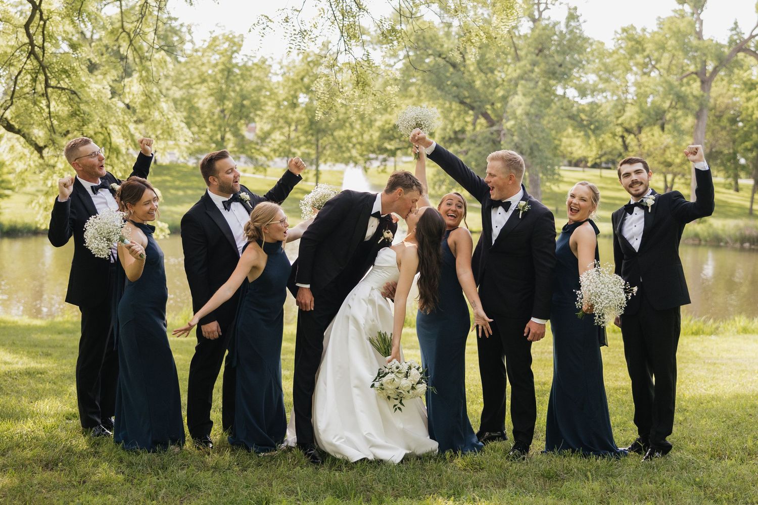 Wedding party celebrates as bride and groom kiss on green lawn, bridesmaids in navy dresses and groomsmen in tuxedos.