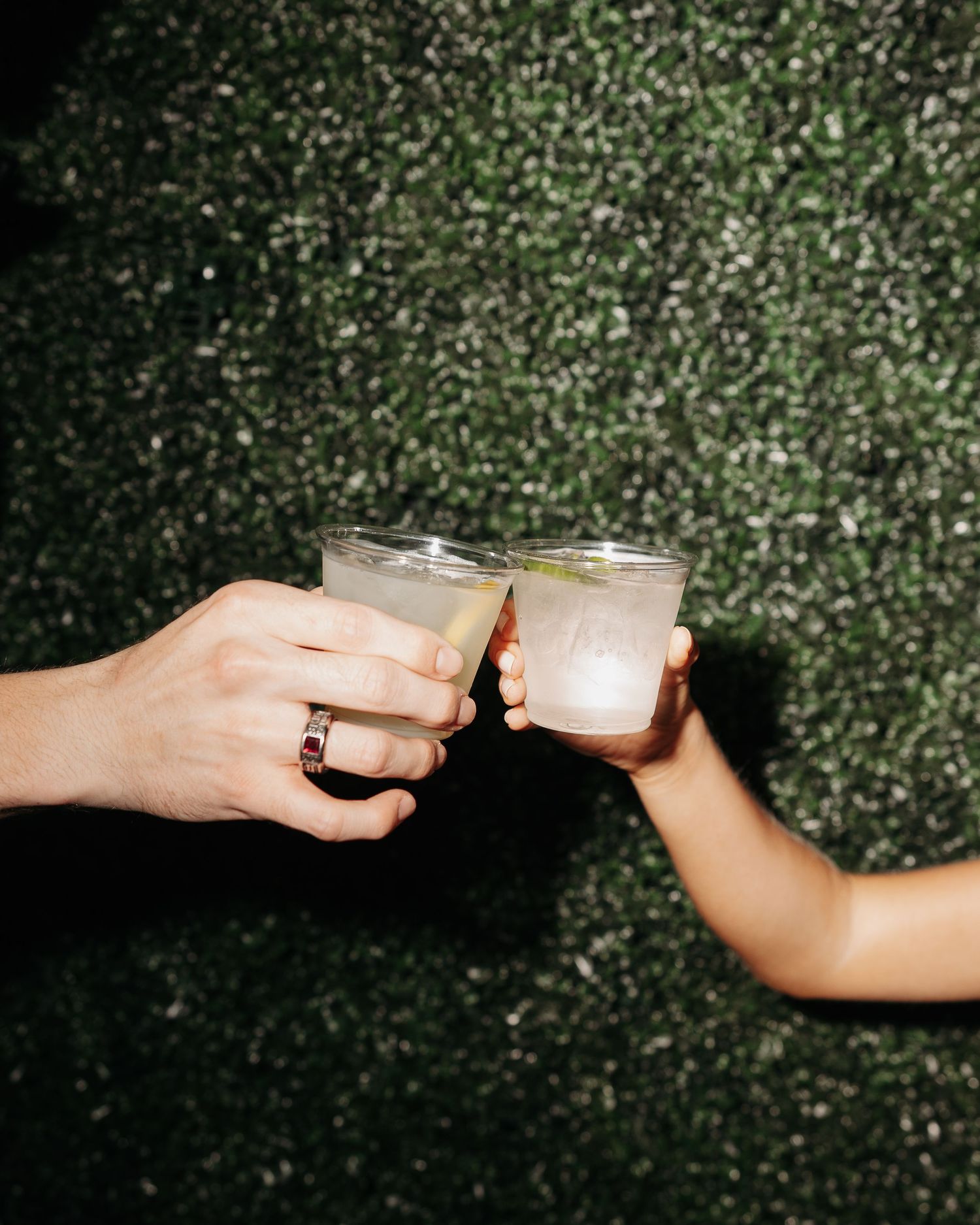 Two hands clinking glasses of margaritas in a toast against a lush green hedge backdrop.