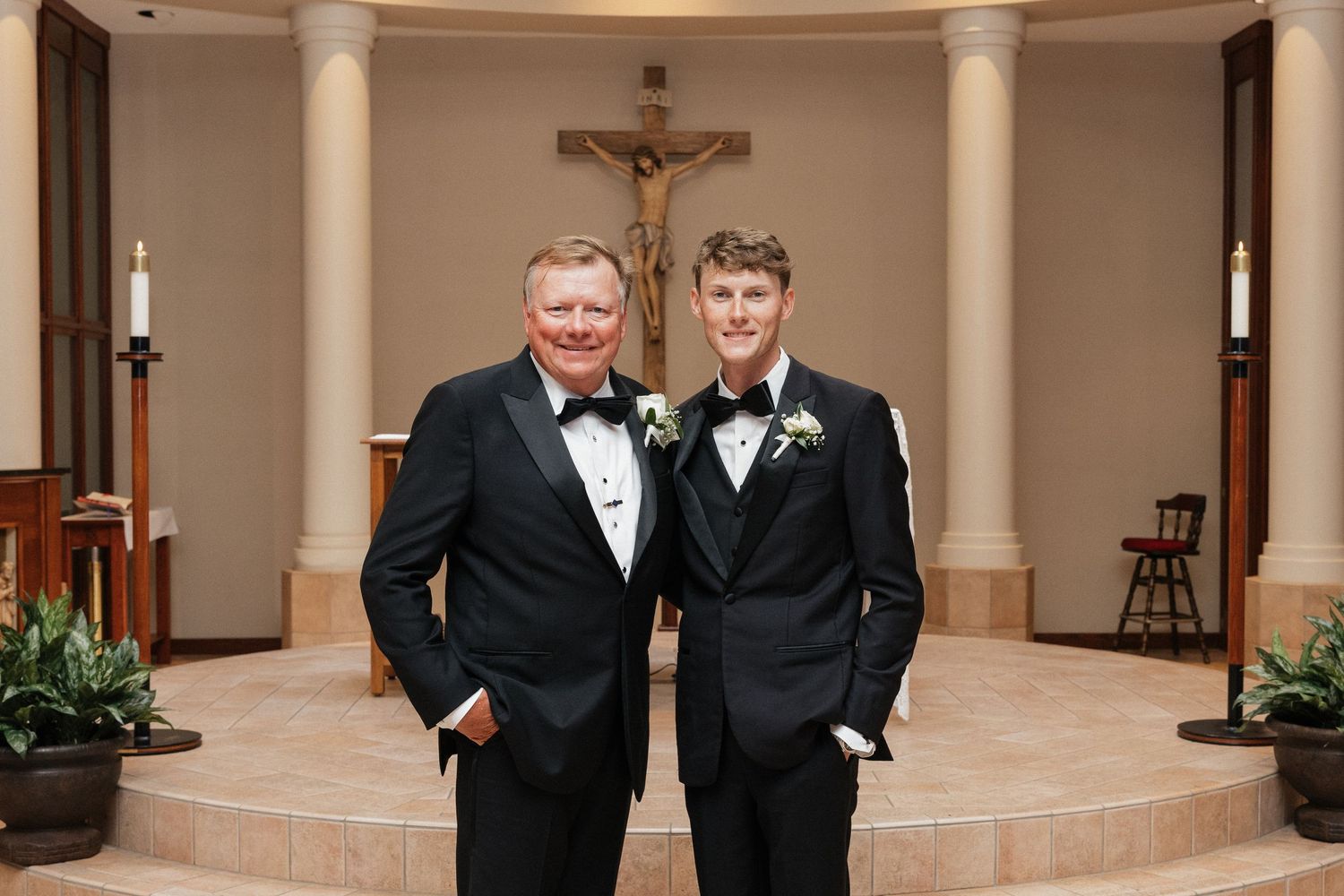 Two men in black tuxedos with boutonnieres pose smiling in a church chapel with a crucifix altar backdrop.
