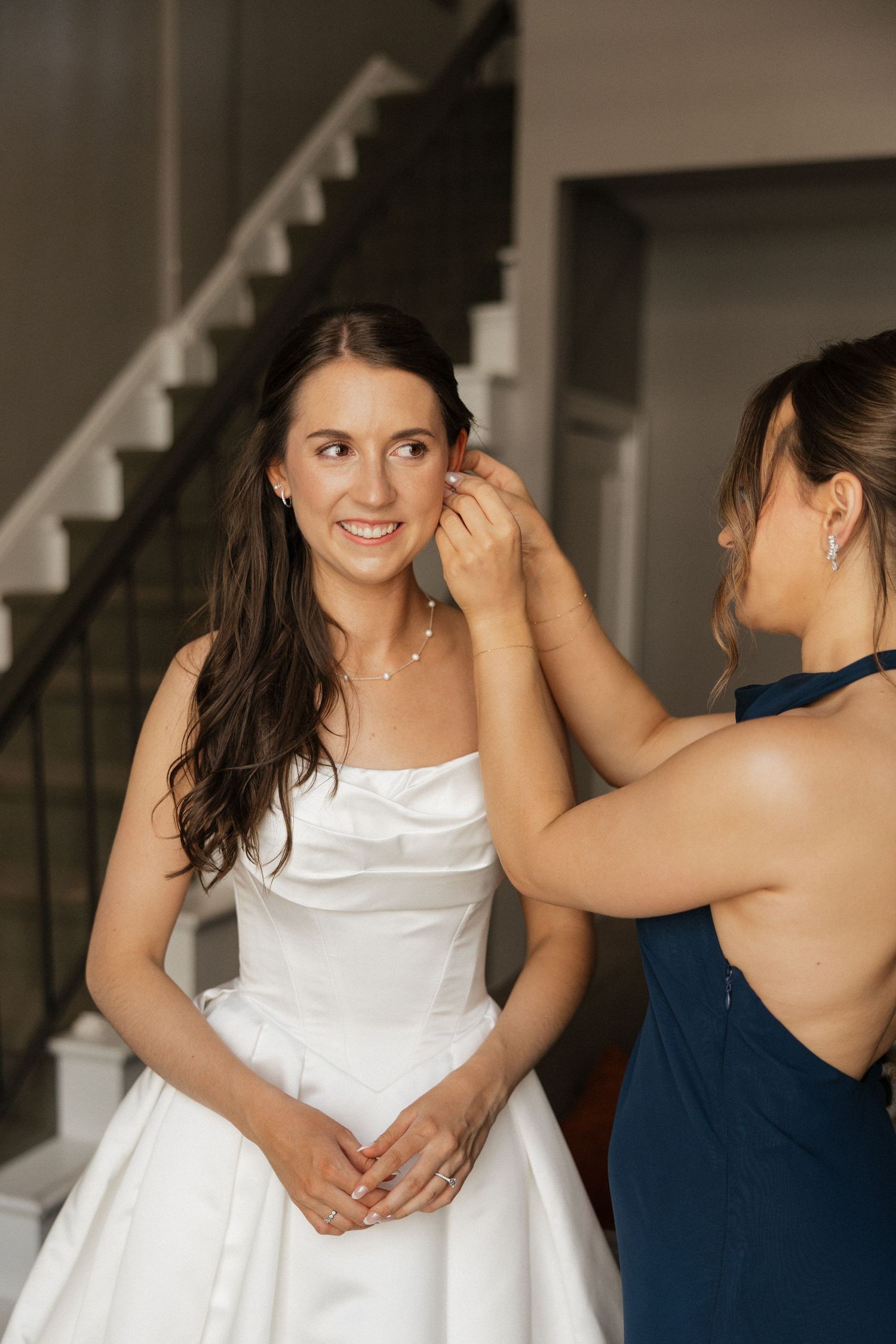 Bride in white strapless gown smiles as bridesmaid in navy dress adjusts her earring near a staircase.