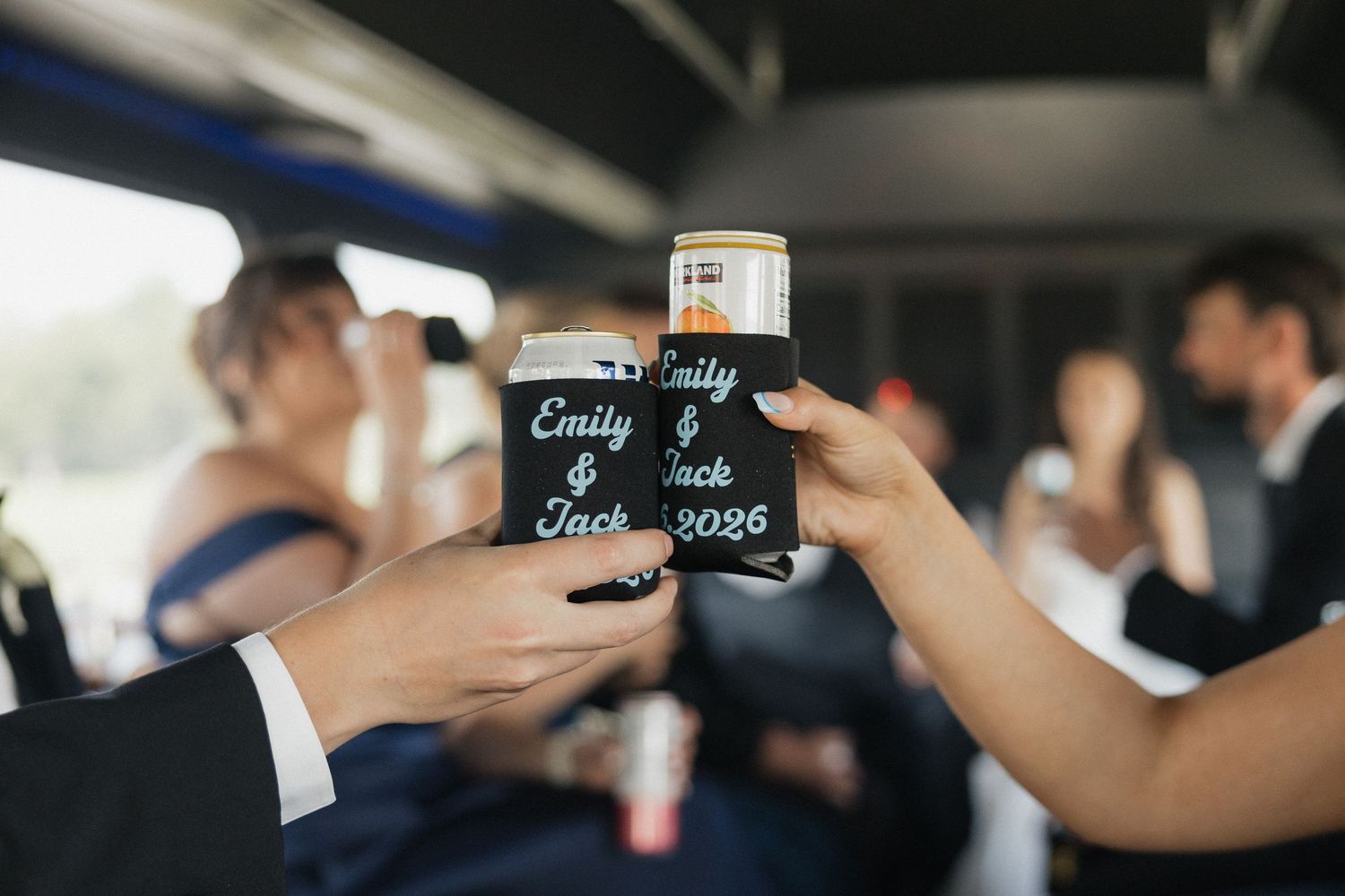 Two black koozies reading Emily & Jack 2026 being toasted together inside a party bus at a wedding.