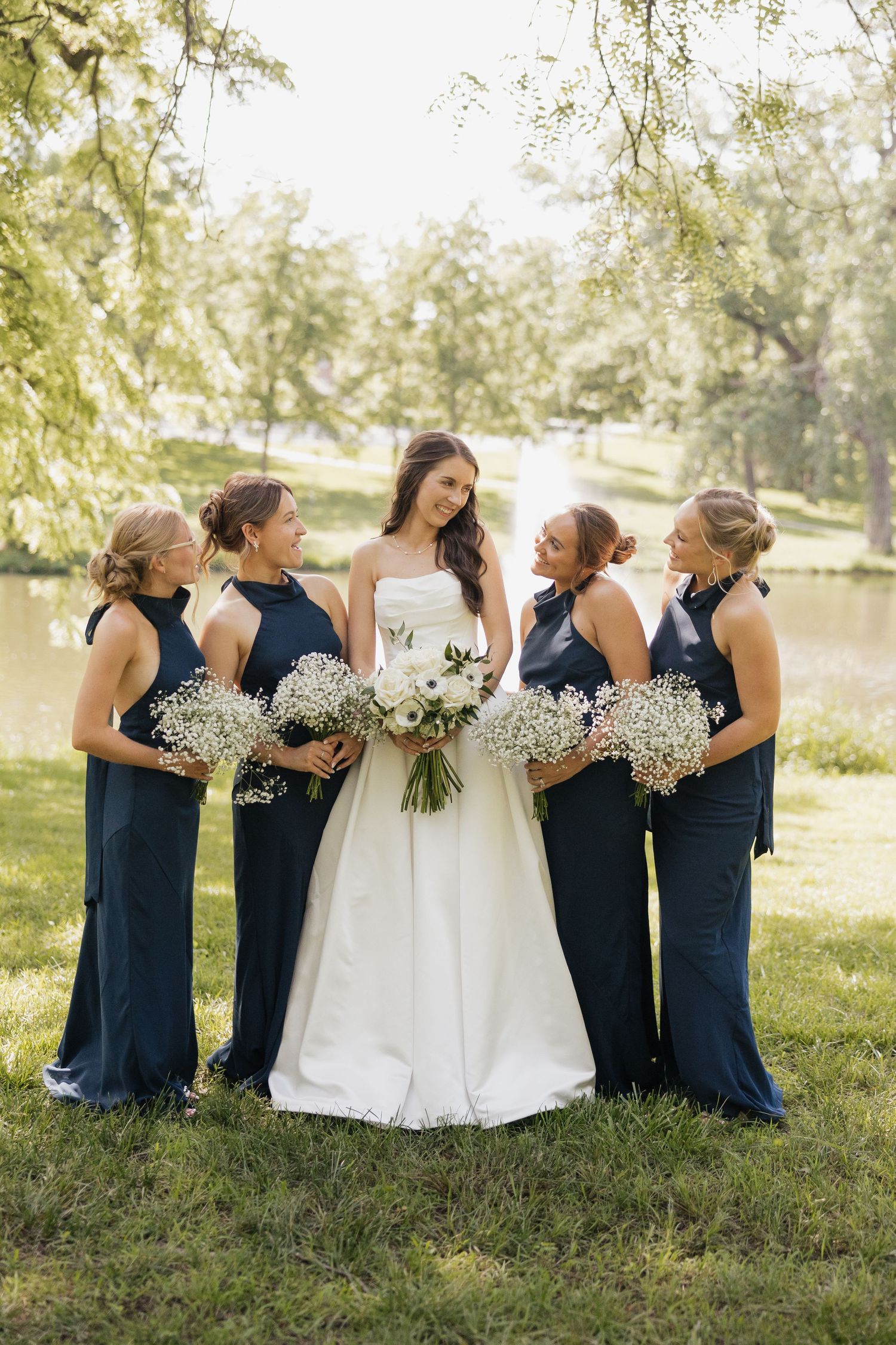 Bride in white gown with four bridesmaids in navy dresses holding baby's breath bouquets outdoors by a pond.
