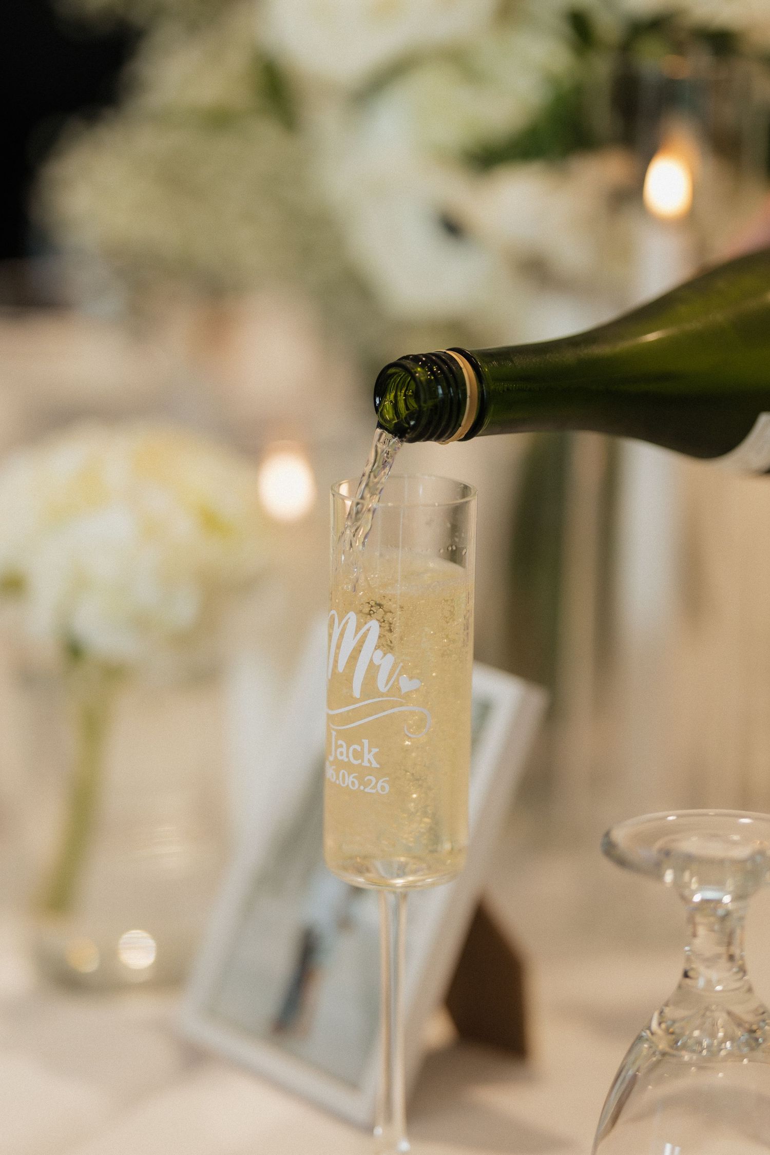 Champagne being poured into a personalized Mr. wedding flute at an elegant reception with white floral arrangements.