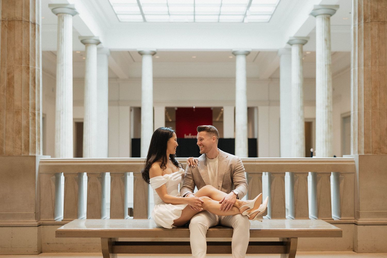 Couple sitting together on a bench in an elegant columned hall, sharing a romantic moment in formal attire.