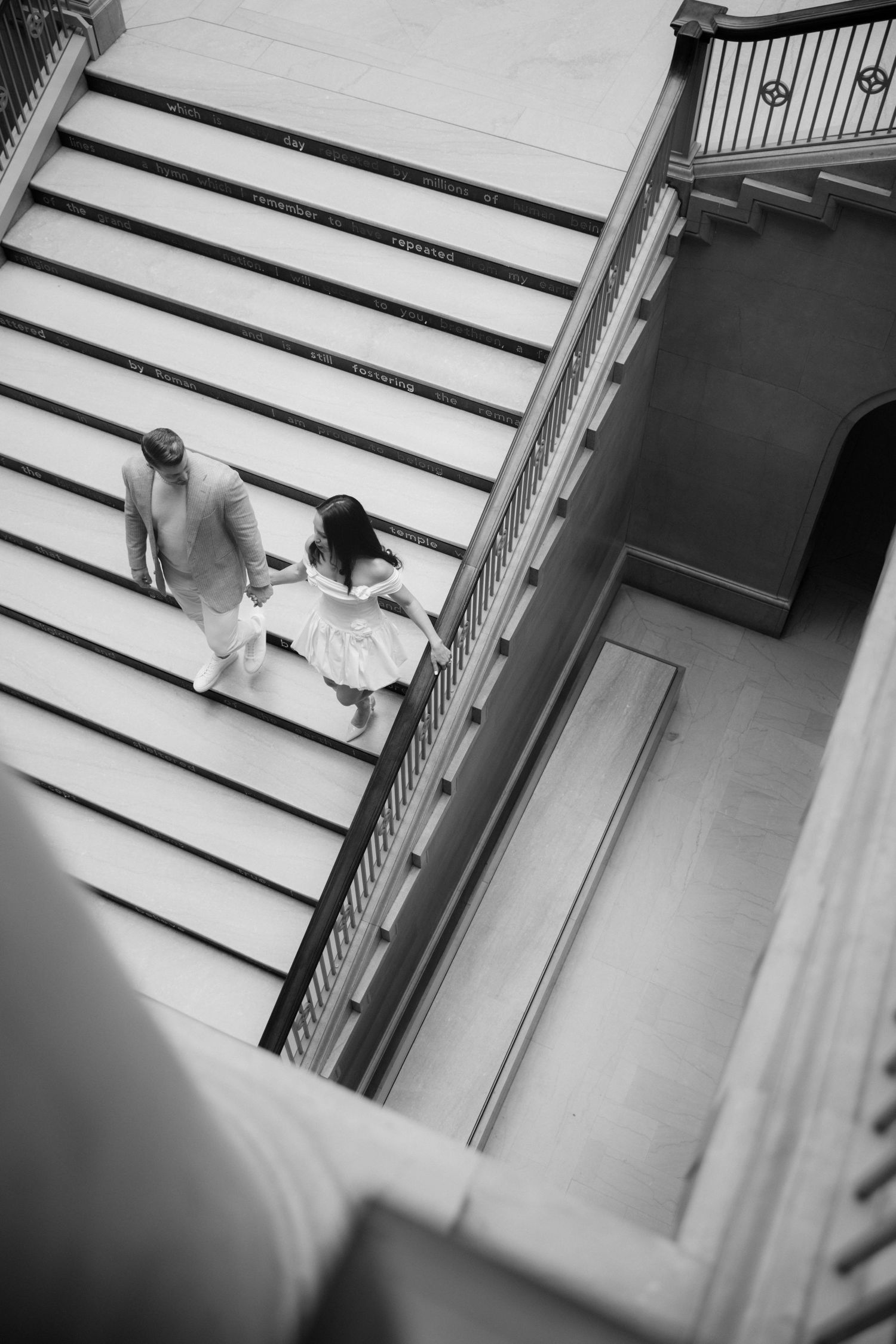 Black and white aerial view of a couple in white attire walking up grand stone staircase in elegant building.