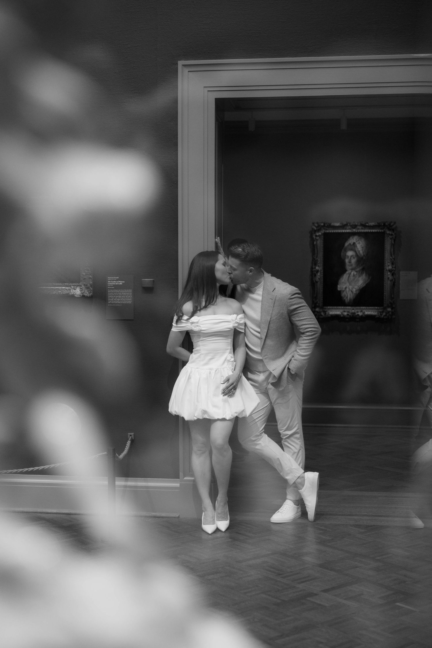 Black and white photo of a couple sharing a kiss in an art museum gallery with a portrait painting in the background.