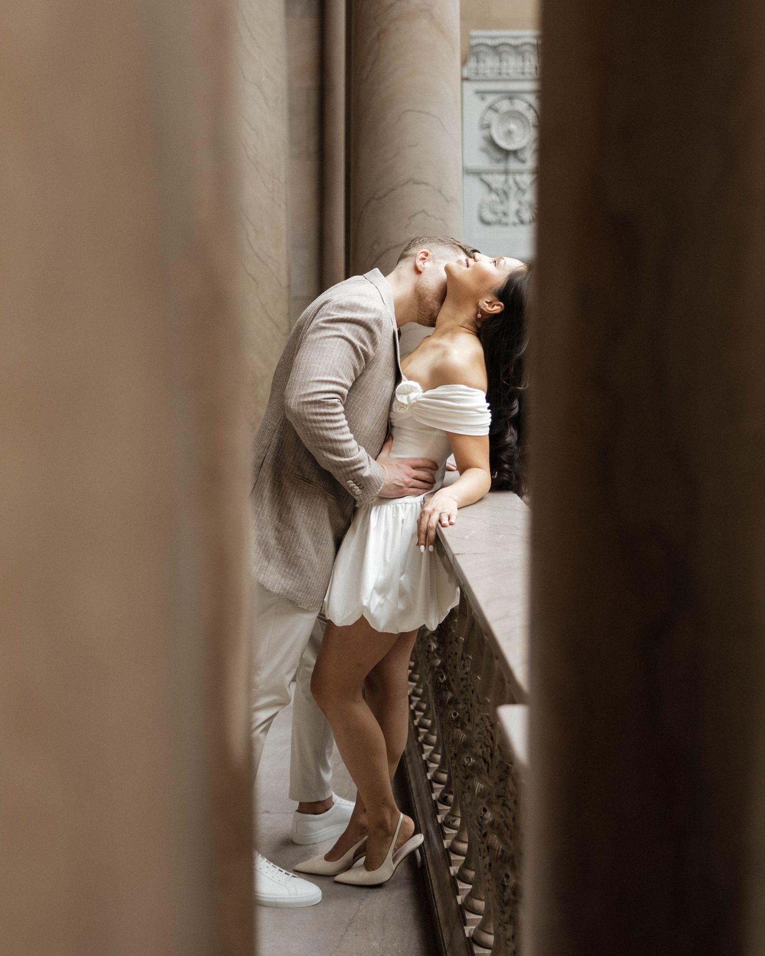 Couple sharing a romantic kiss in an elegant architectural setting, man in beige suit, woman in white off-shoulder mini dress.