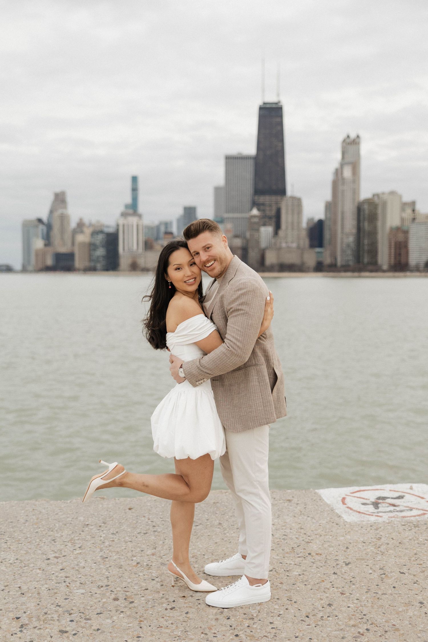 Couple embracing and smiling by Lake Michigan with Chicago skyline backdrop, woman in white dress, man in tan suit.