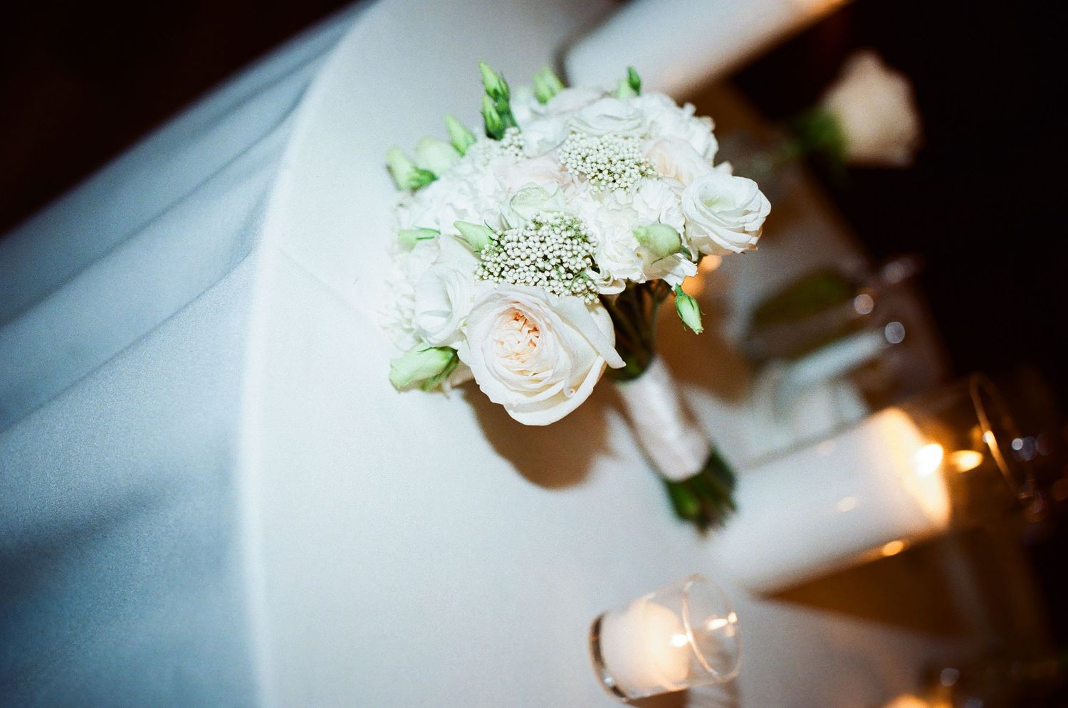 White rose wedding bouquet with baby's breath displayed on a table with candles at an elegant wedding reception.