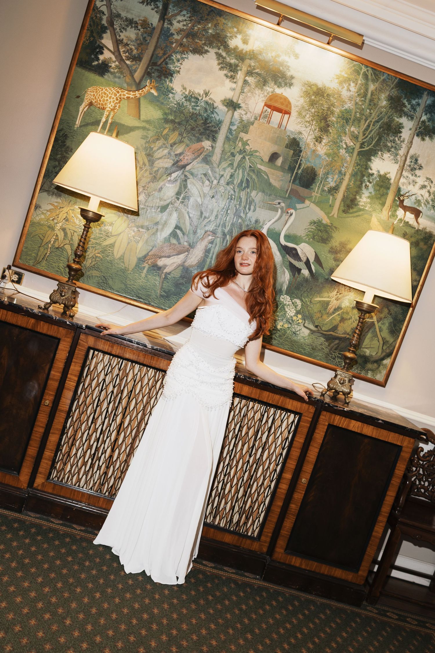 A red-haired bride in a white gown poses before a tropical mural flanked by gold lamps on a dark wood sideboard.