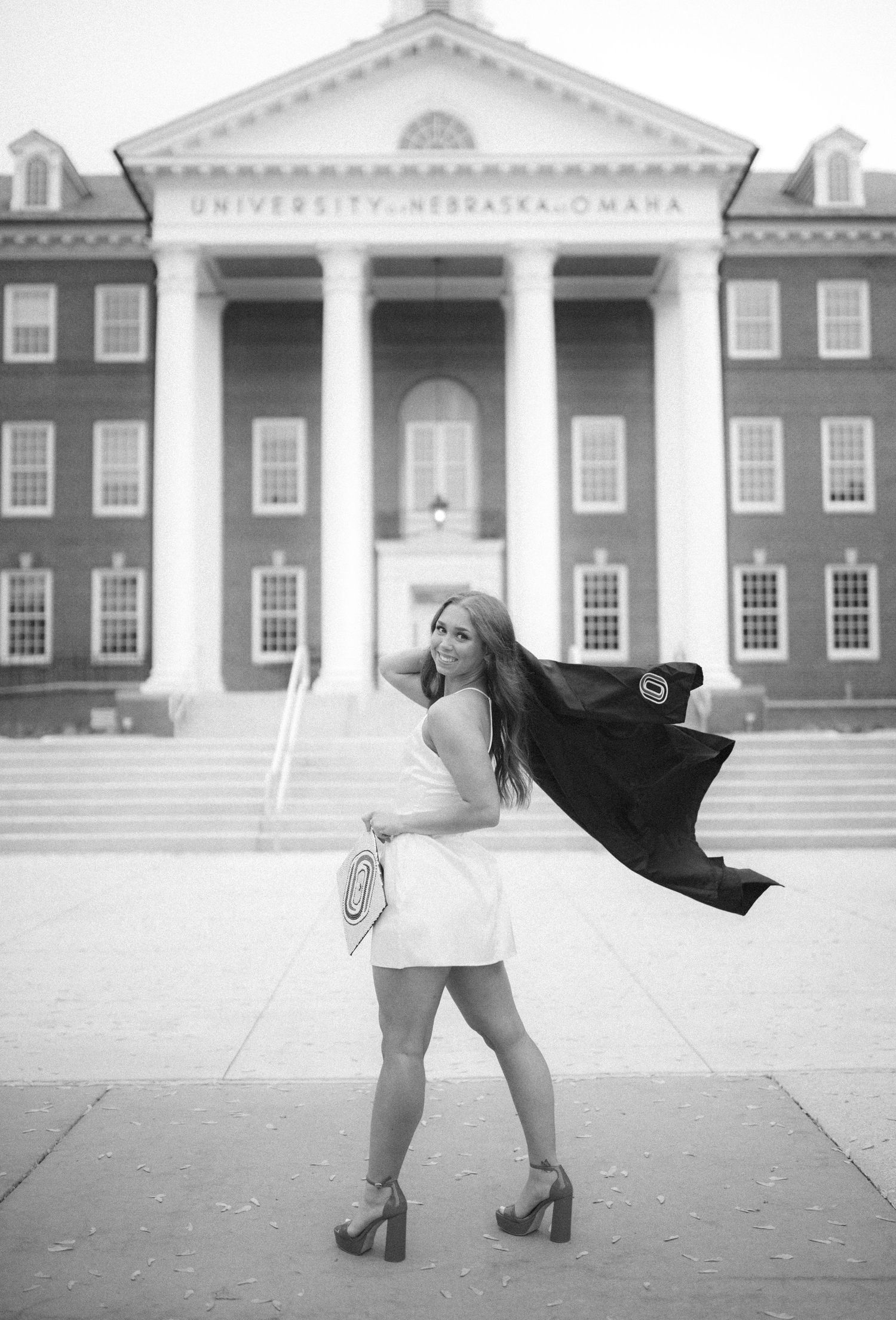 A graduate poses in a flowing black graduation gown outside the University of Nebraska-Omaha building in black and white.