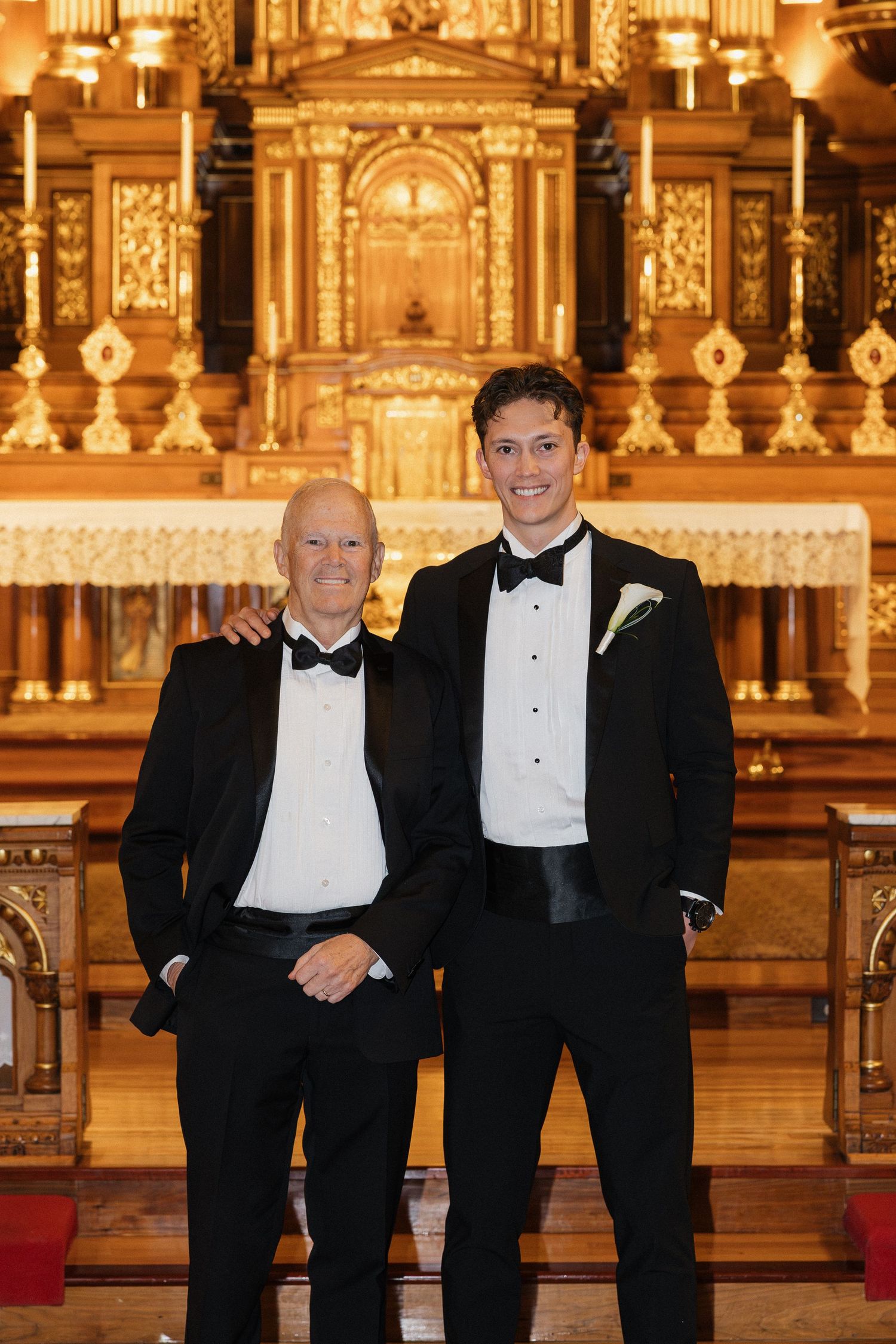 Two men in formal black tuxedos stand together in front of an ornate golden church altar.