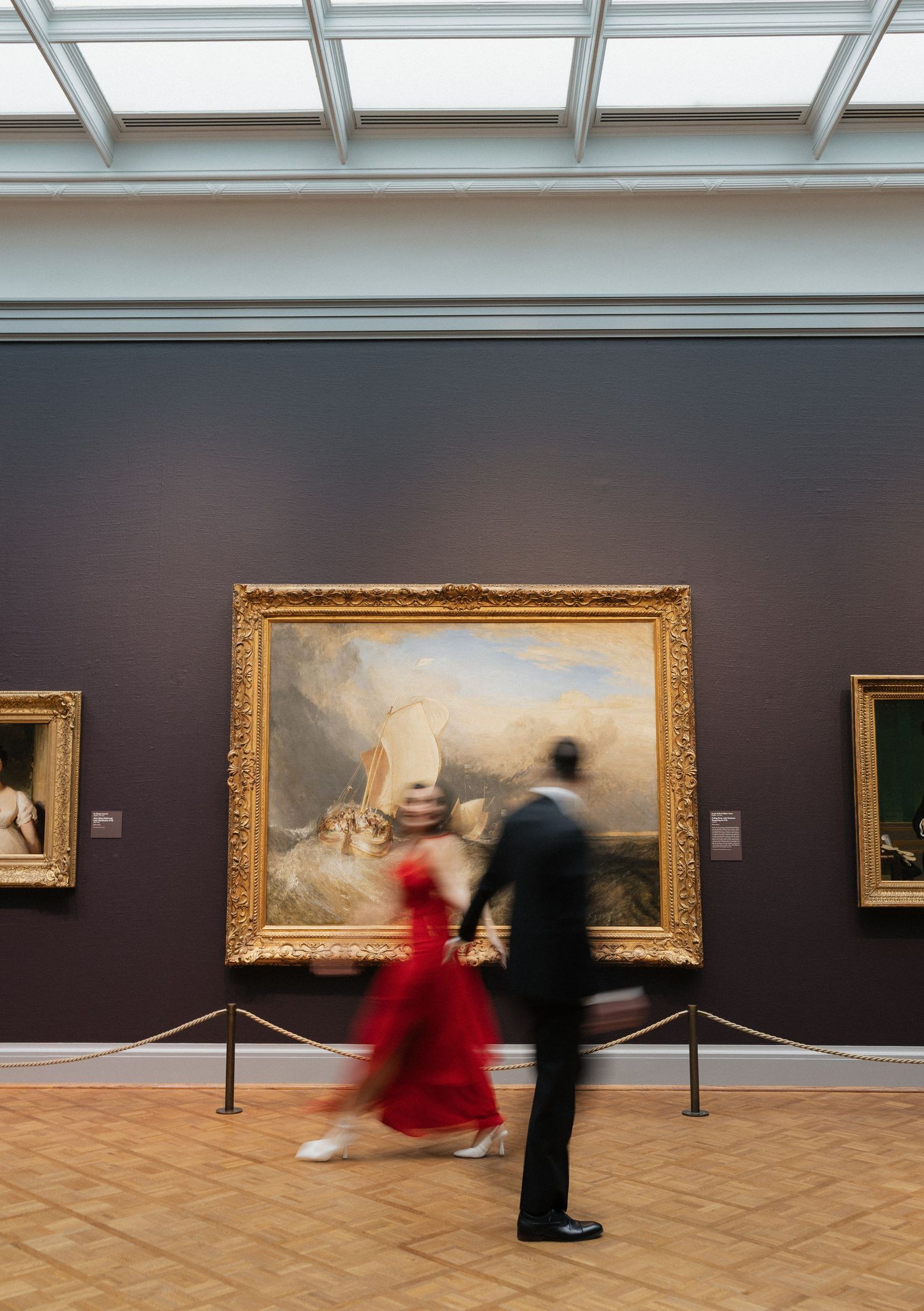 Two visitors in motion blur view a classical painting in an ornate gold frame at an art gallery with skylights above.