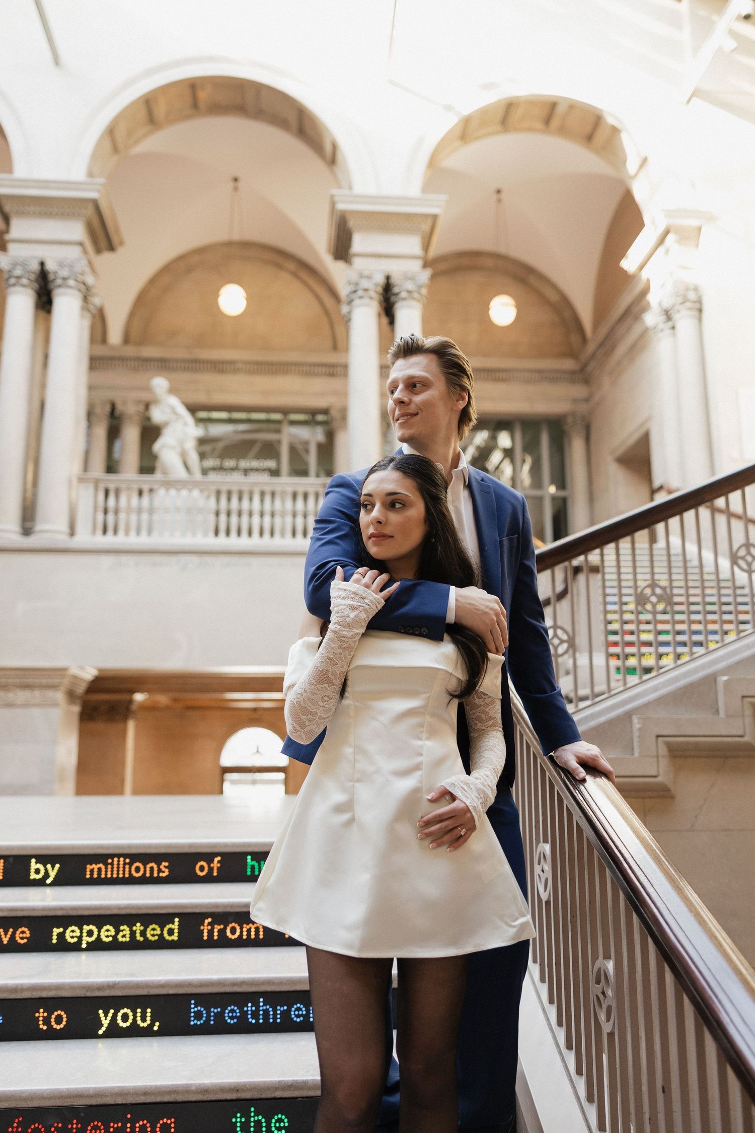 Romantic couple posing on grand marble staircase with classical architecture.