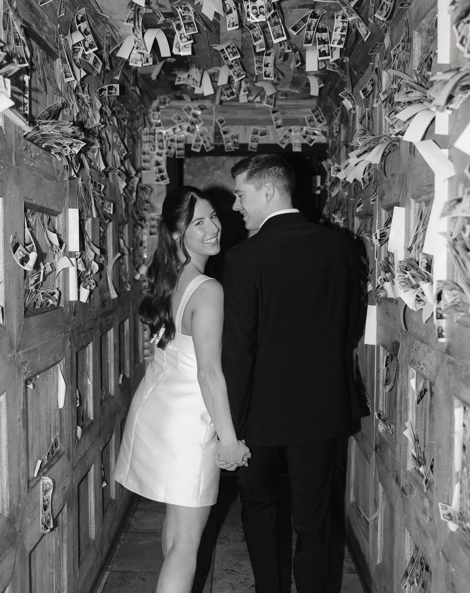 A black and white photograph shows guests mingling in a dimly lit hallway decorated with hanging vines and foliage.
