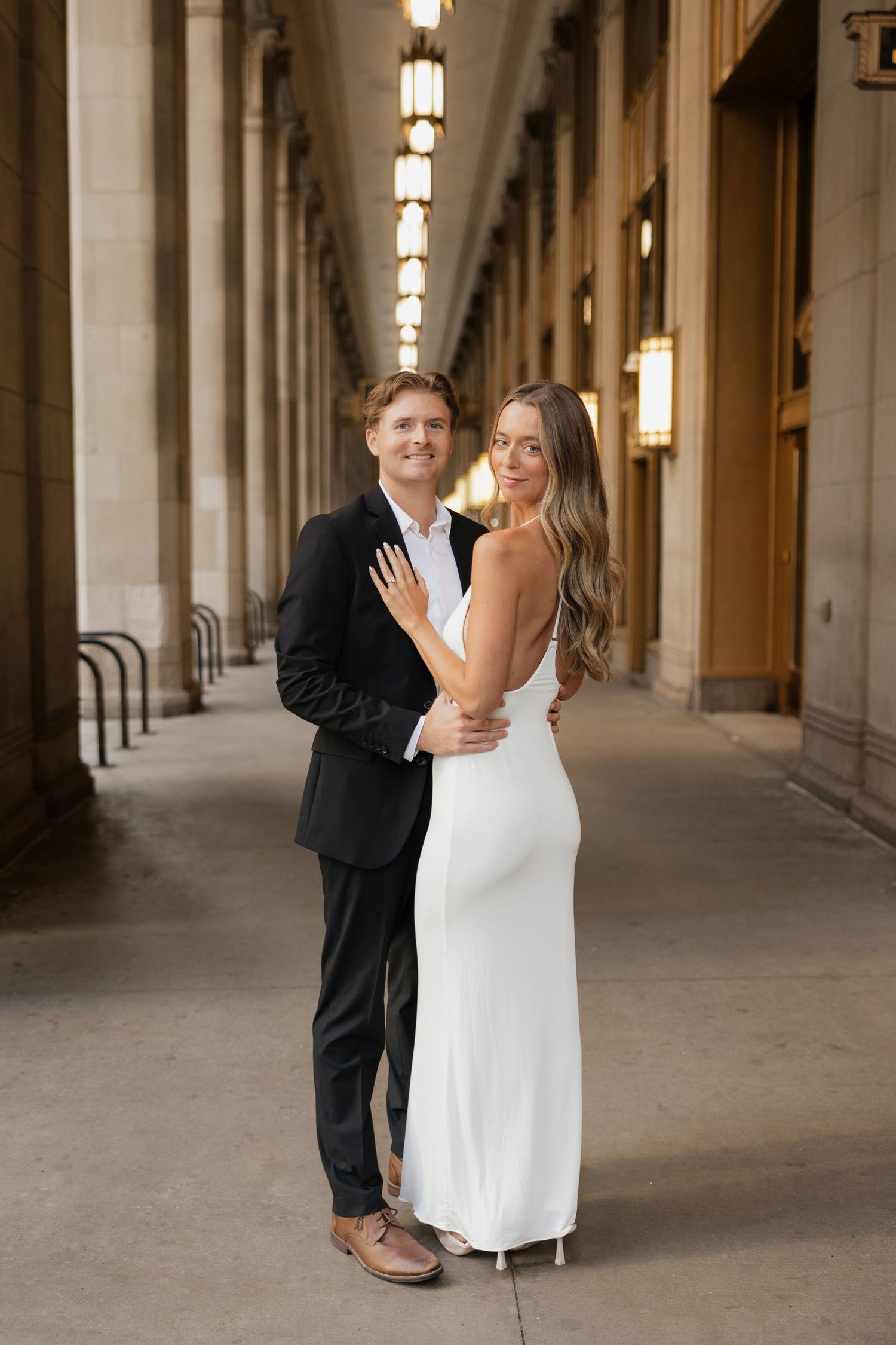 Engaged couple posing in elegant arcade hallway, man in black suit embracing woman in white gown.