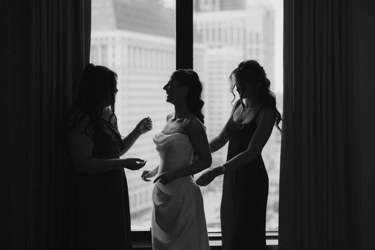 Black and white silhouette of bride being helped into wedding dress by two bridesmaids in front of bright window.