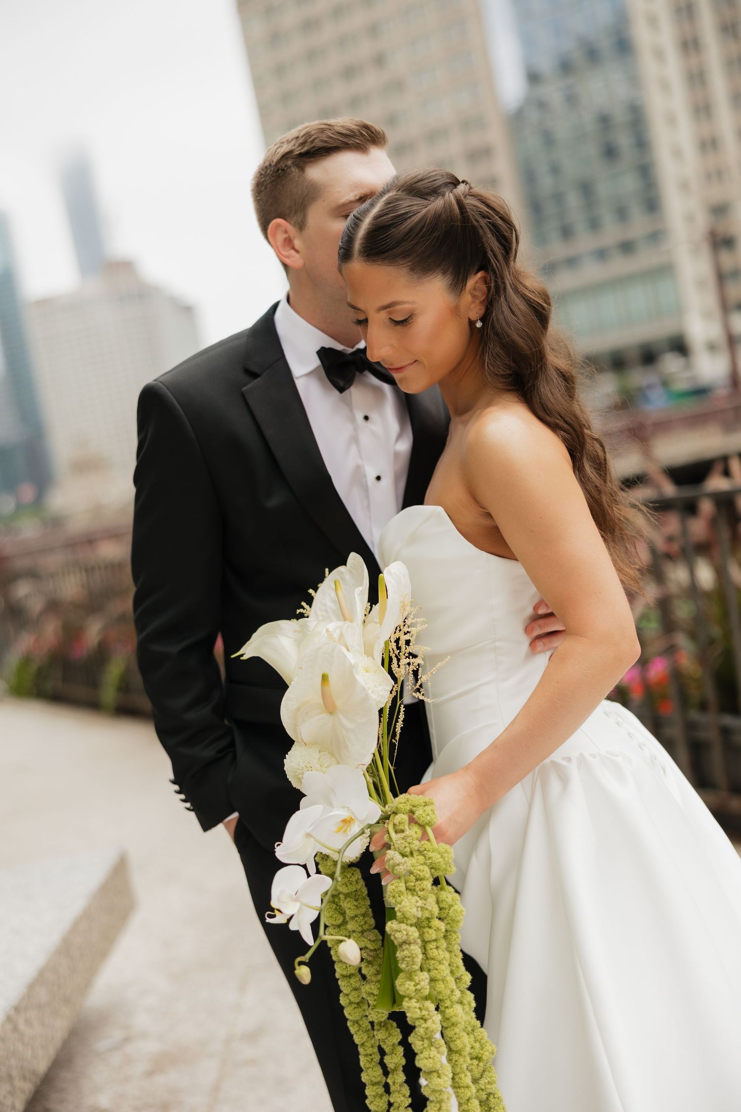 Bride and groom share an intimate moment outdoors, she holds a white floral bouquet with green accents, city skyline behind them.